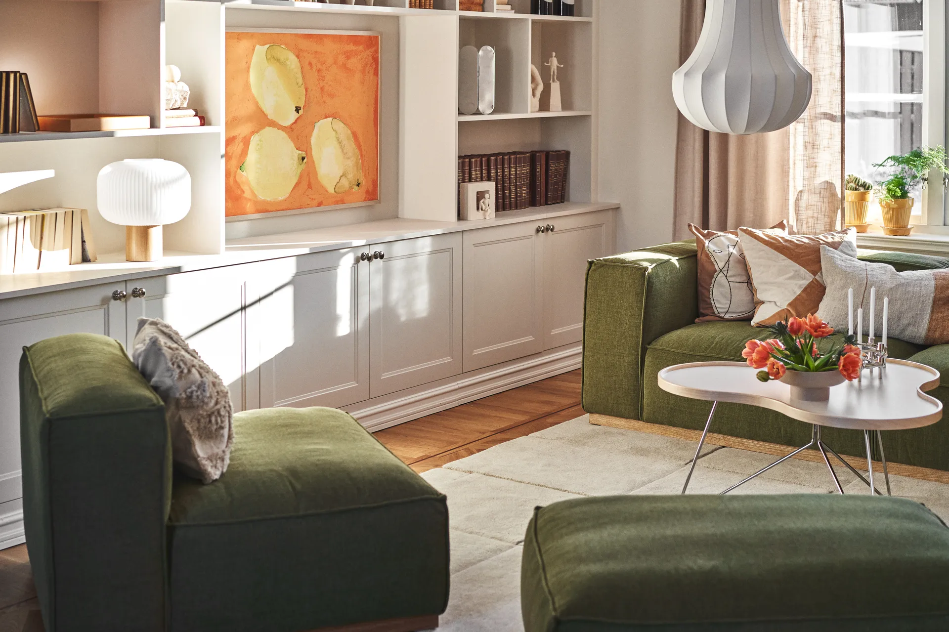 Sonnenhelles Wohnzimmer mit grünen Sofas, weißem Couchtisch, orangen Tulpen, Kerzen und weißen Einbauregalen mit Kunst.