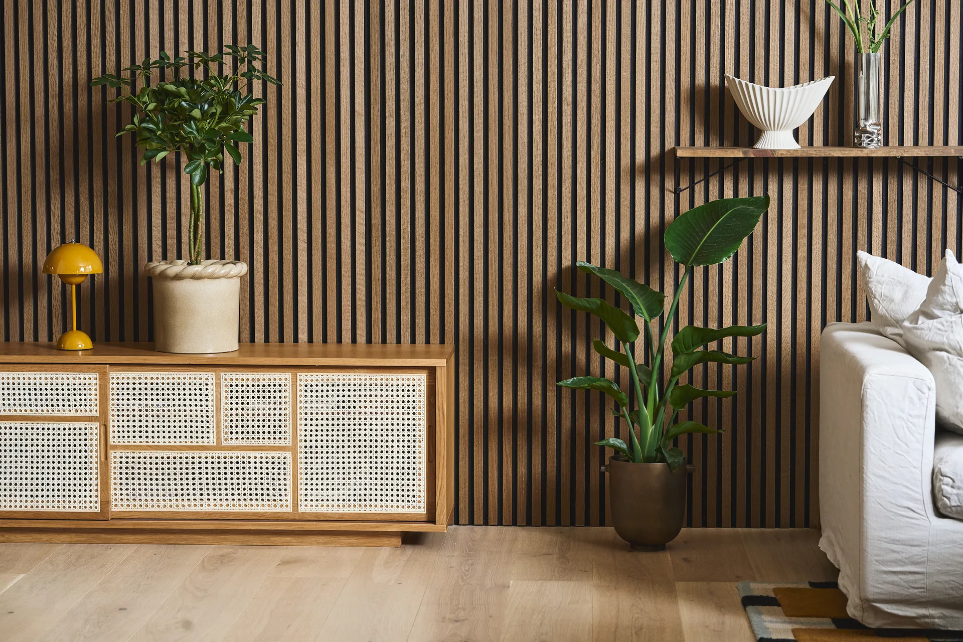 Modernes Wohnzimmer mit Lattenholzwand, Rattanschrank, gelber Lampe, Topfpflanzen und einem weißen Sofa.