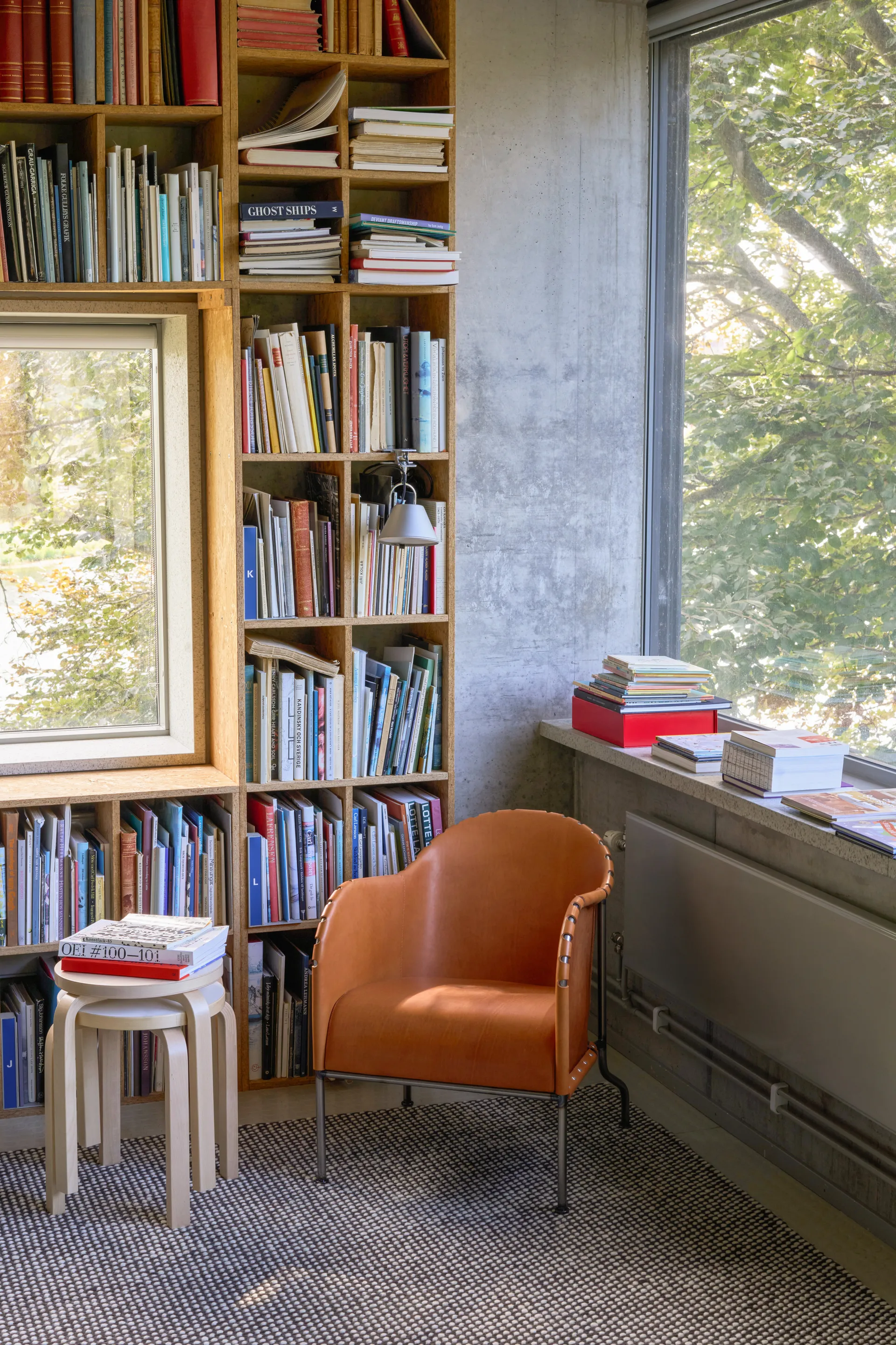 Gemütlicher Raum mit großer Holz-Bücherwand, hellbraunem Ledersessel, kleinem Hocker mit Büchern und Fenster mit Baumblick.