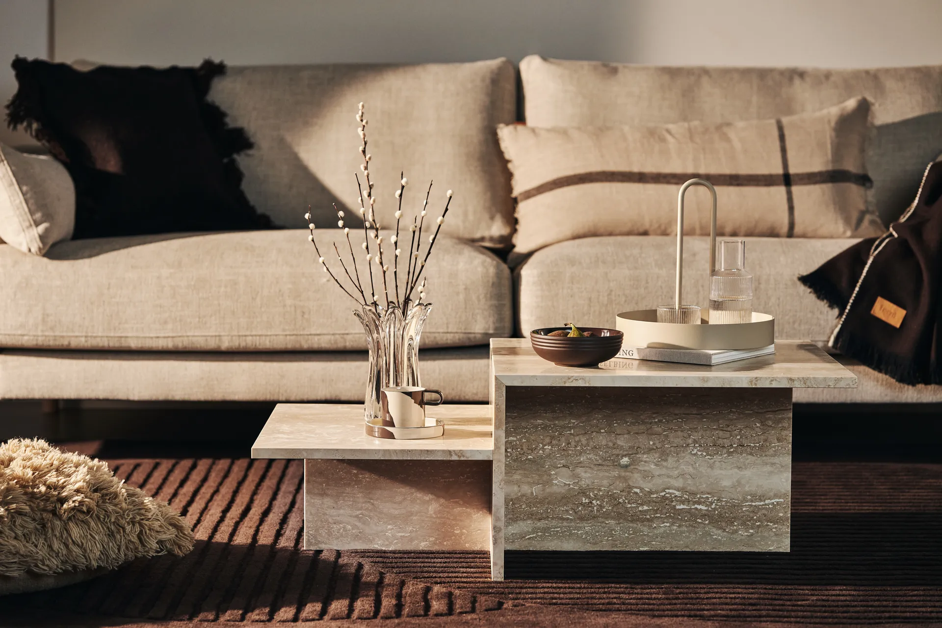 Gestufter Couchtisch aus beige Stein vor einem neutralen Sofa mit dekorativen Accessoires.
