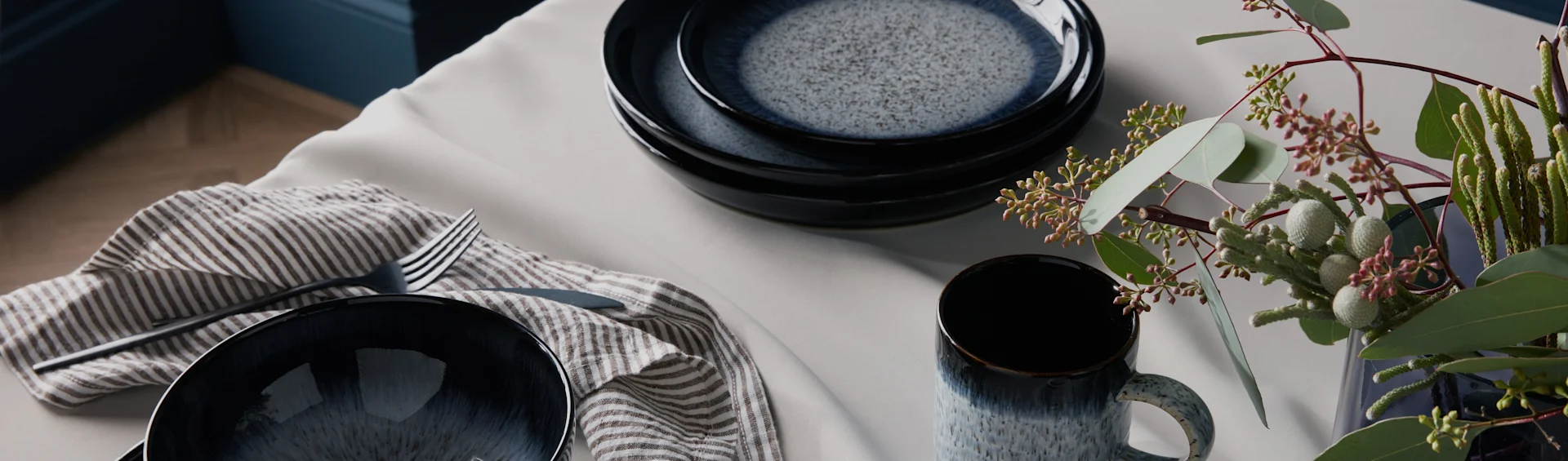 Gesprengtes dunkelblaues Keramikgeschirr, gestreifte Serviette, Besteck und grünes Laub auf einem hellen Tisch.