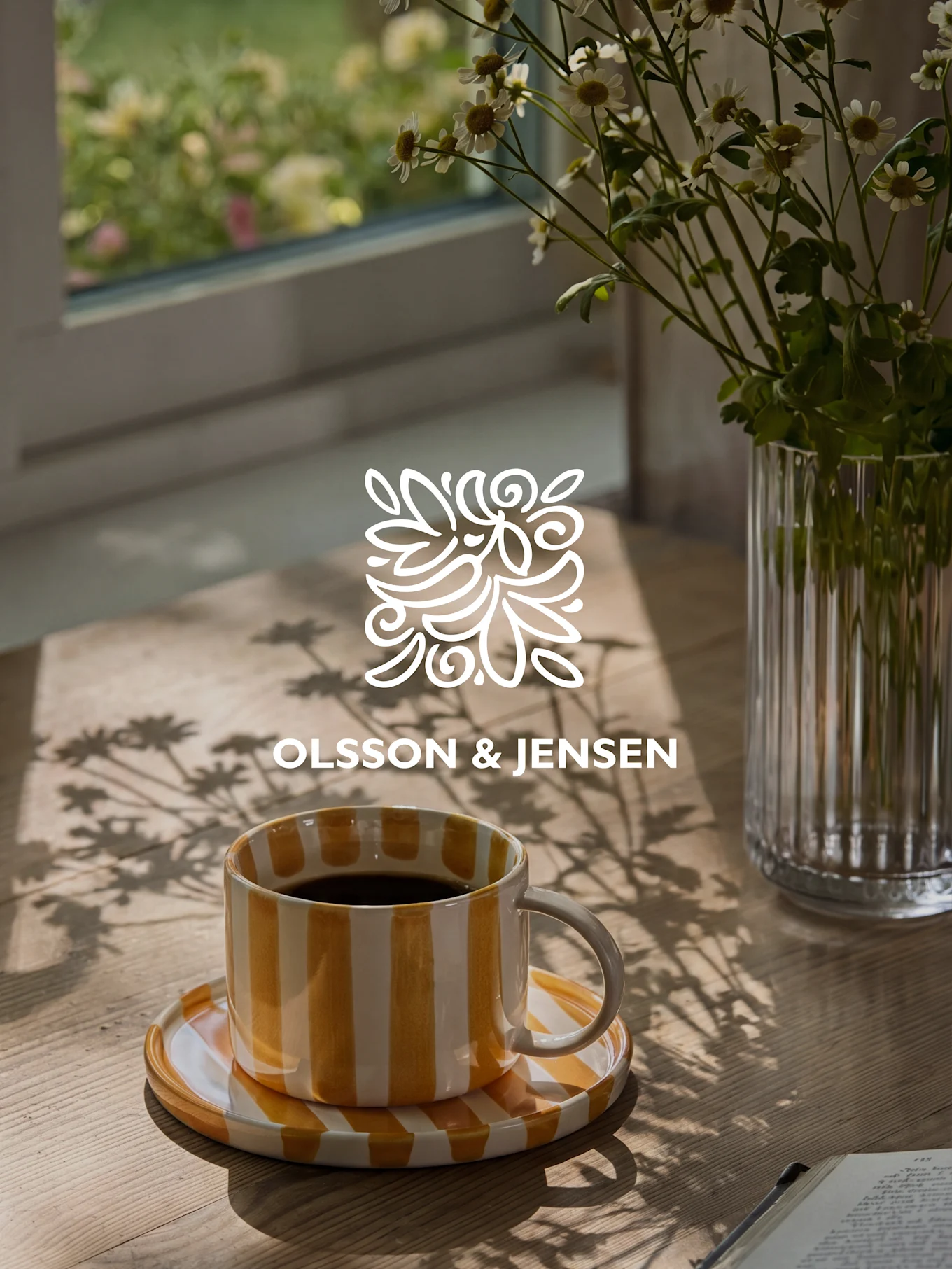 Gelb-weiß gestreifte Kaffeetasse auf einem Holztisch mit einer Vase weißer Blumen und Sonnenlichtschatten, Olsson & Jensen Logo.