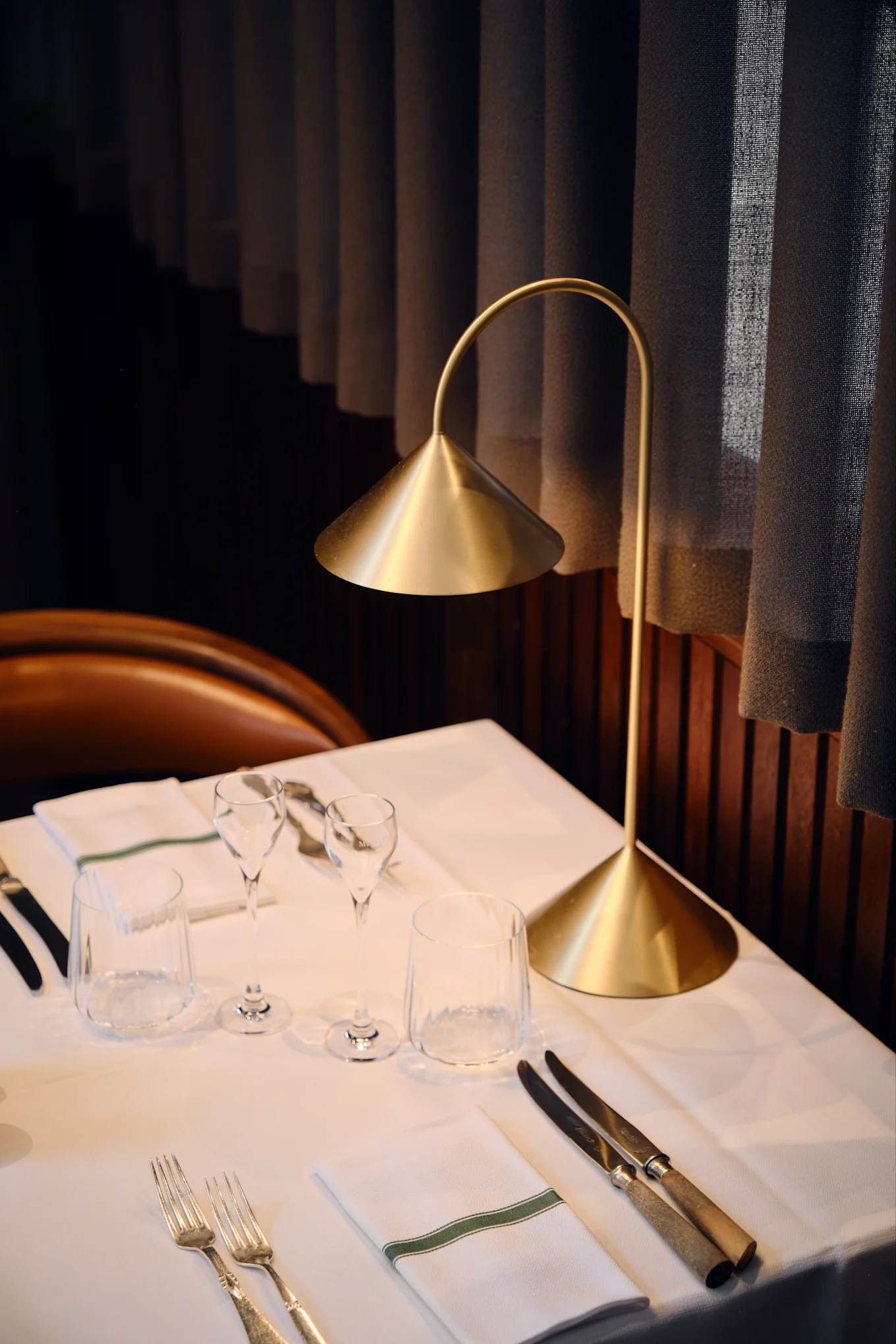 Eine moderne goldene Tischlampe beleuchtet einen gedeckten Restauranttisch mit weißer Wäsche, Gläsern, Besteck und einer Serviette mit grünem Streifen.