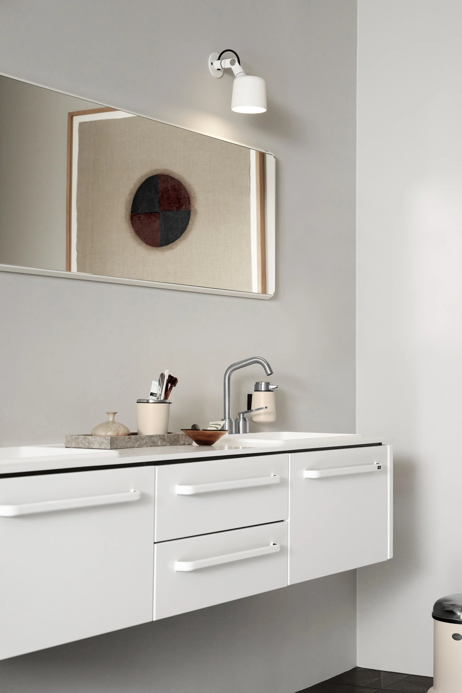 Modernes Badezimmer mit weißem Waschtisch, silberner Armatur, cremefarbenem Seifenspender, Spiegel mit abstrakter Kunst und weißer Wandleuchte.