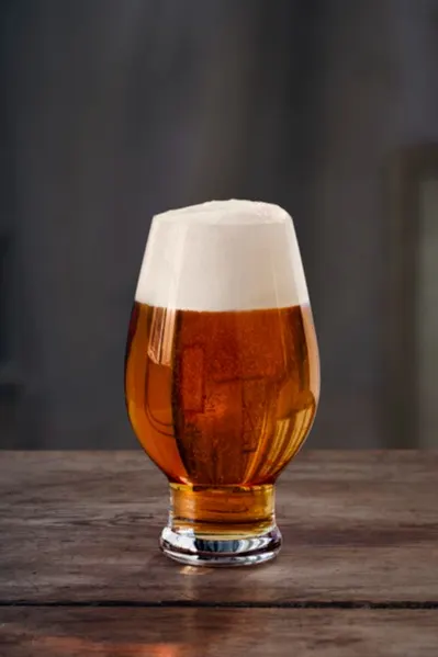 Tulpenförmiges Glas mit bernsteinfarbenem Bier und dickem weißem Schaum auf einem rustikalen Holztisch.
