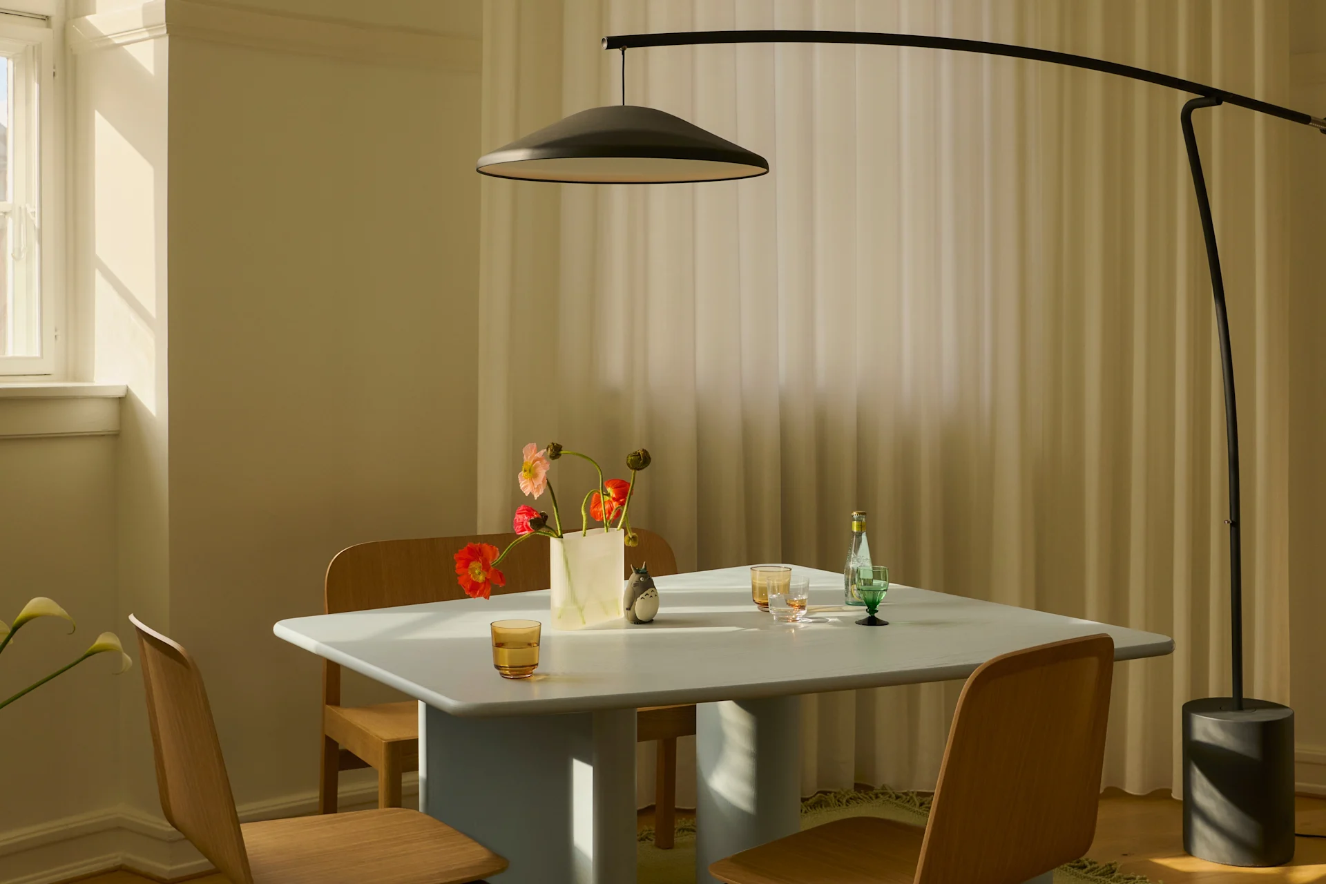 Ein modernes Esszimmer mit hellblauem Tisch, Holzstühlen, schwarzer Bogen-Stehleuchte und einer Vase bunter Blumen im Sonnenlicht.