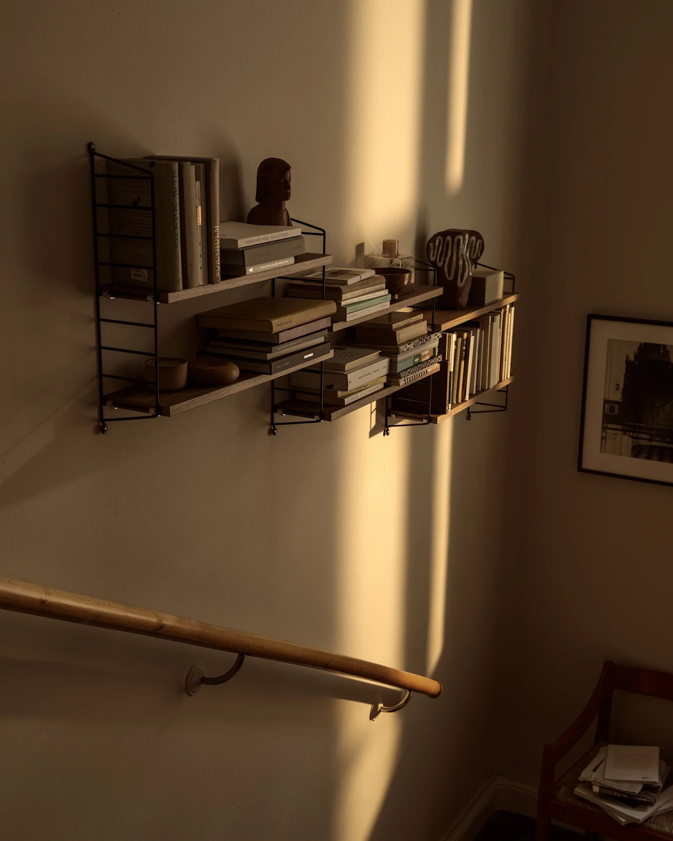 Wandregale mit Büchern und Deko, gebadet in warmem Sonnenlicht mit starken vertikalen Schatten über einem Holzhandlauf.