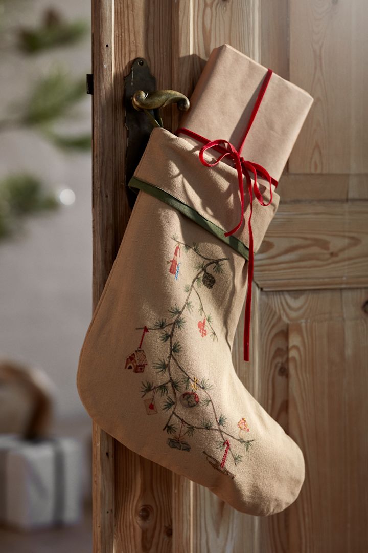 Die Weihnachtsdeko für 2025 sind von der Natur inspiriert, wie dieser Strumpf von Kähler, den Sie hier an einer Holztür vor einem Weihnachtsbaum und verpackten Geschenken hängen sehen. 