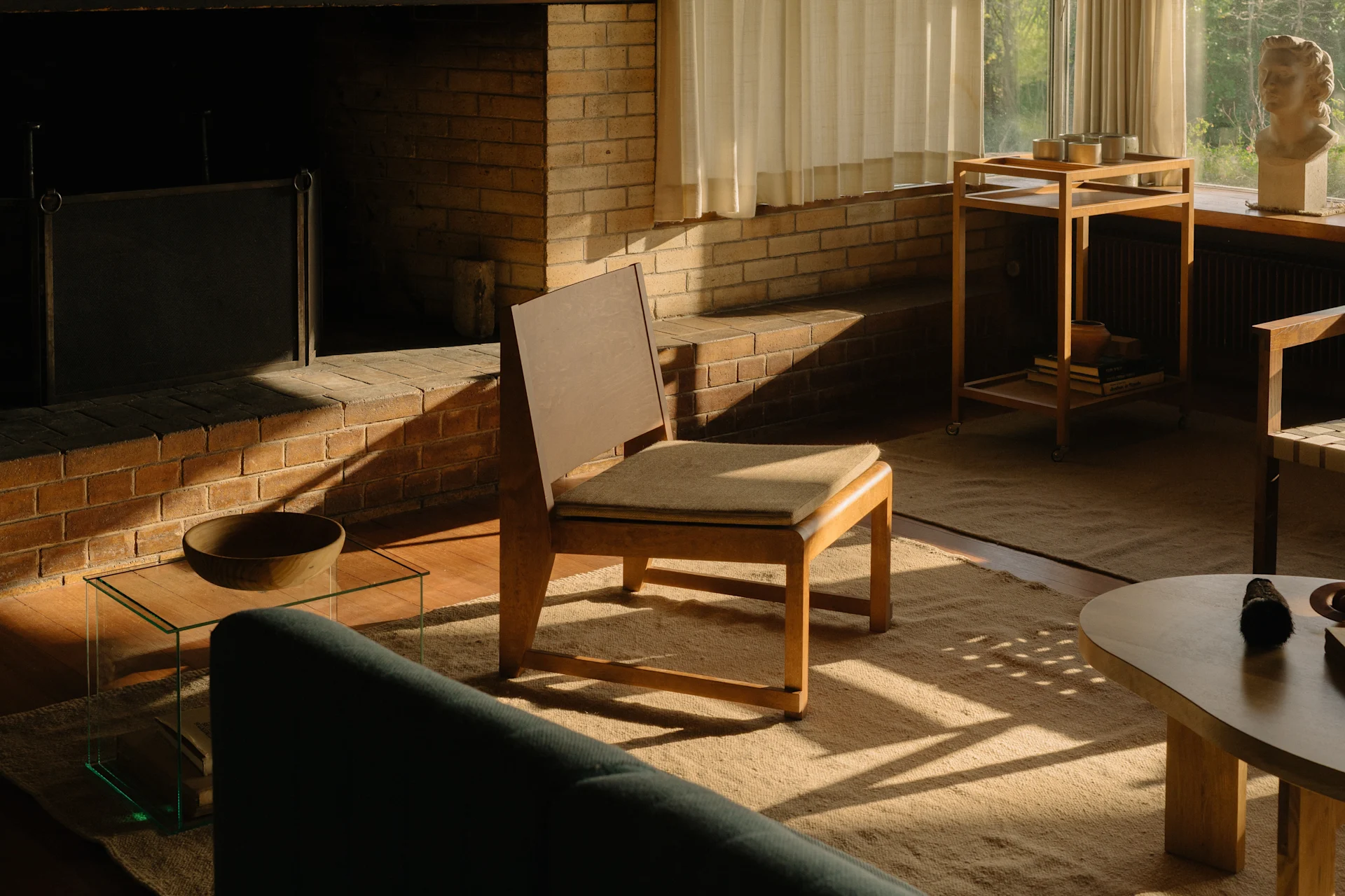 Ein sonnenbeschienenes Wohnzimmer mit Ziegelkamin, Holzstuhl und grünem Sofa, mit einer Büste am Fenster.