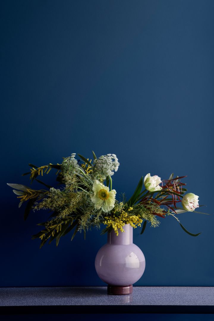 Runde lila Vase von Broste Copenhagen mit Frühlingsblumen vor blauer Wand - zwei Farben, welche die Wohntrends 2022 dominieren.