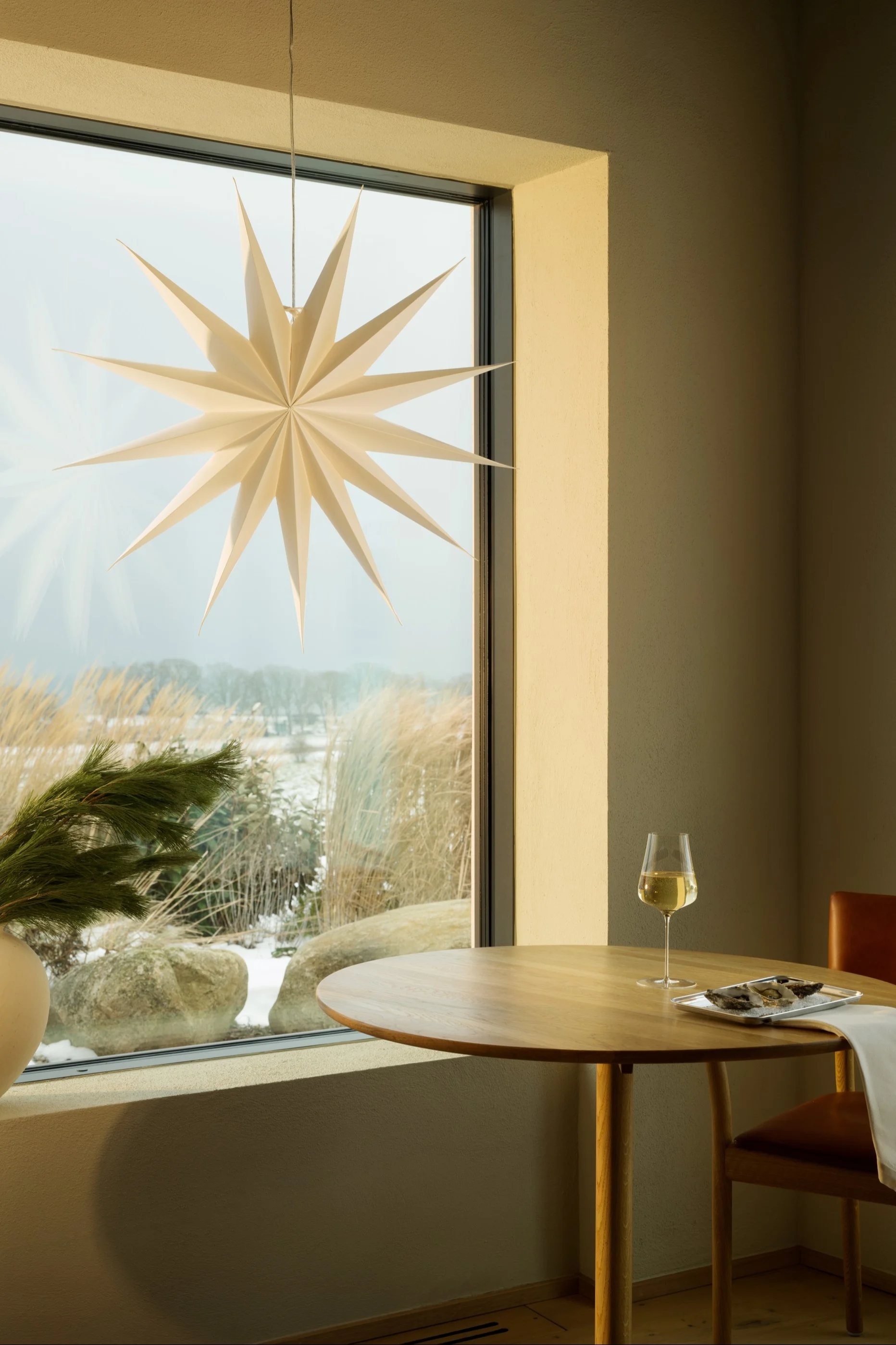 Helles Fenster mit beigen Vorhängen, immergrüner Topfpflanze und weißer Sternlaterne. Helle Bettwäsche vorn.