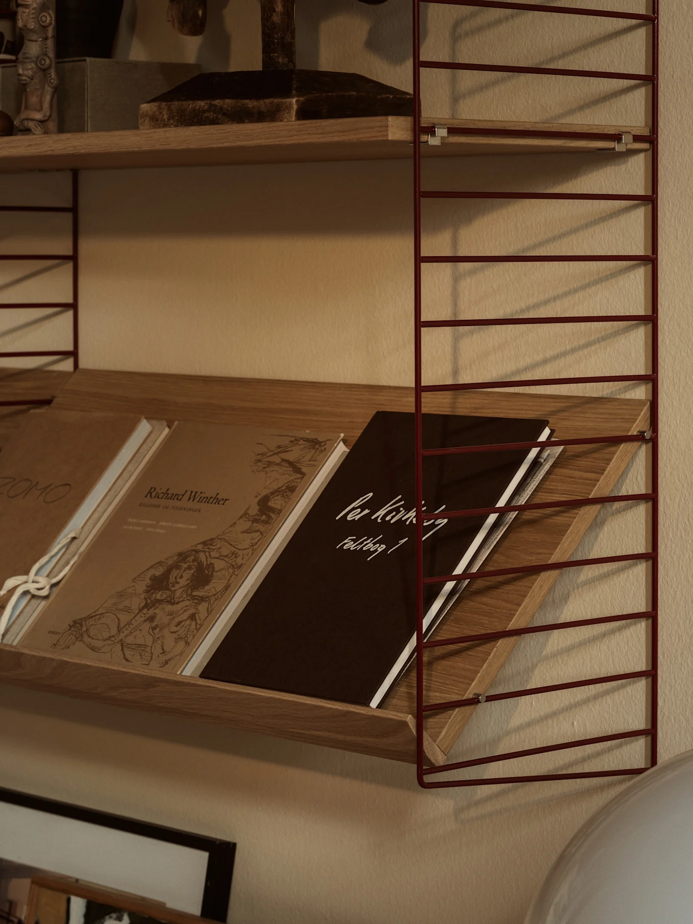 Ein rotes Metallregalsystem mit Holzbrettern. Das untere Regalbrett hält schräg gestellte Bücher, darunter ein dunkelbraunes mit 'Per Kirkeby Feldbog 1'.