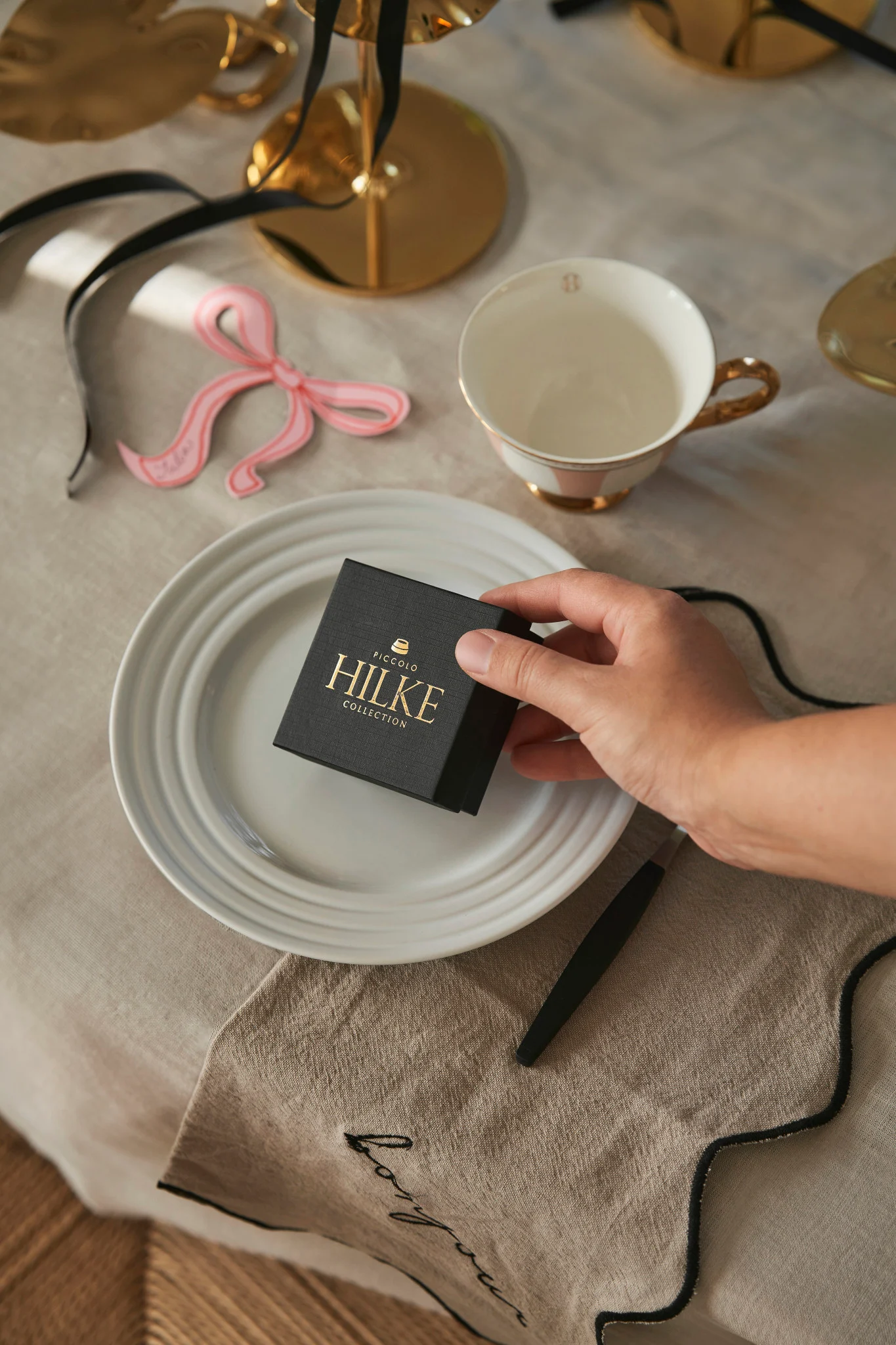 Feiern Sie den Galentine's Day zu Hause mit Ihren Lieblingsmenschen. Hier sehen Sie eine Hand, die eine Schachtel von Hilke Collection als kleines Geschenk für die Gäste auf ein Gedeck stellt.