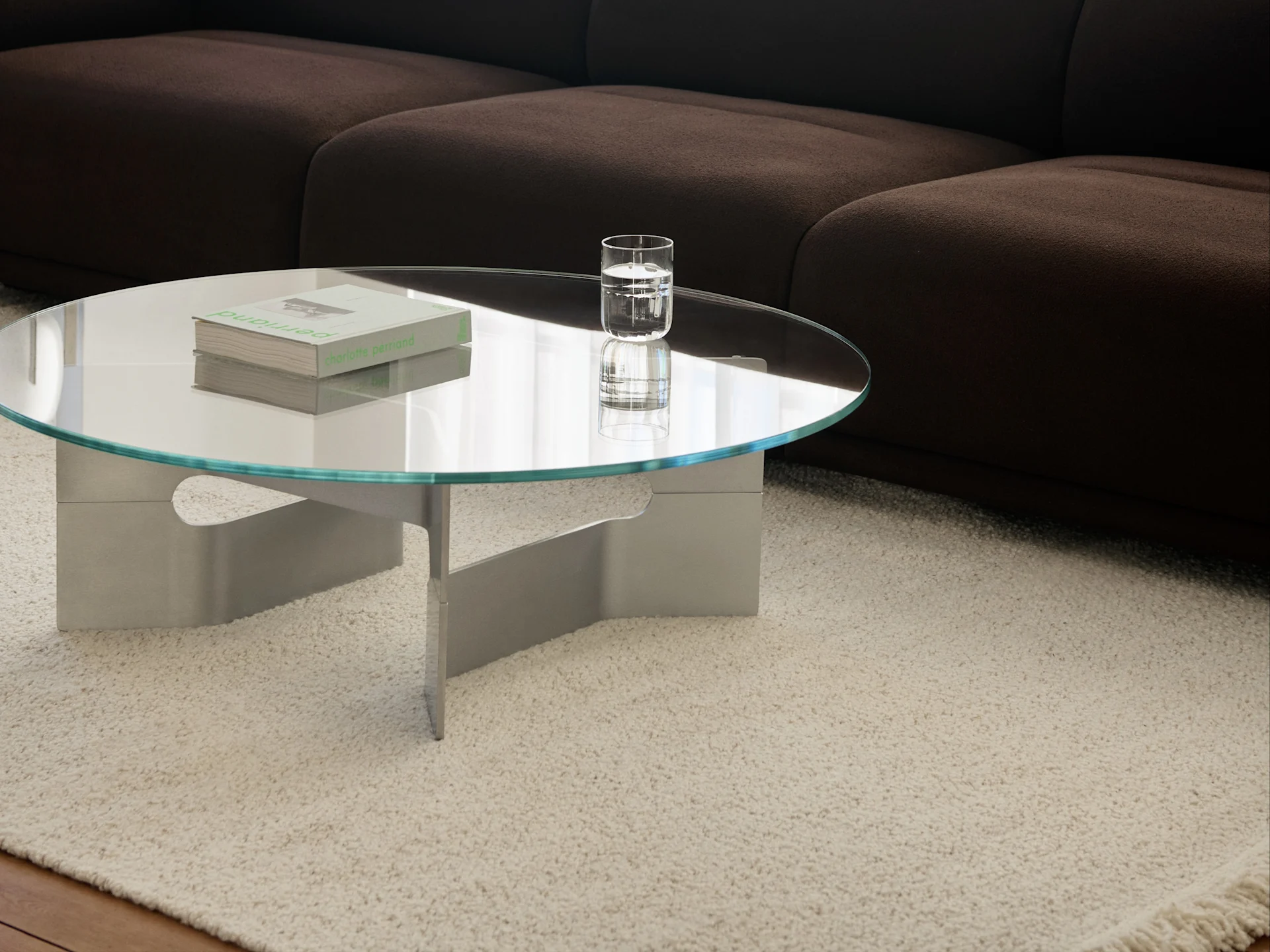 Runder Couchtisch aus Glas mit Büchern und einem Glas Wasser auf einem cremefarbenen Zottelteppich vor einem dunkelbraunen Sofa.