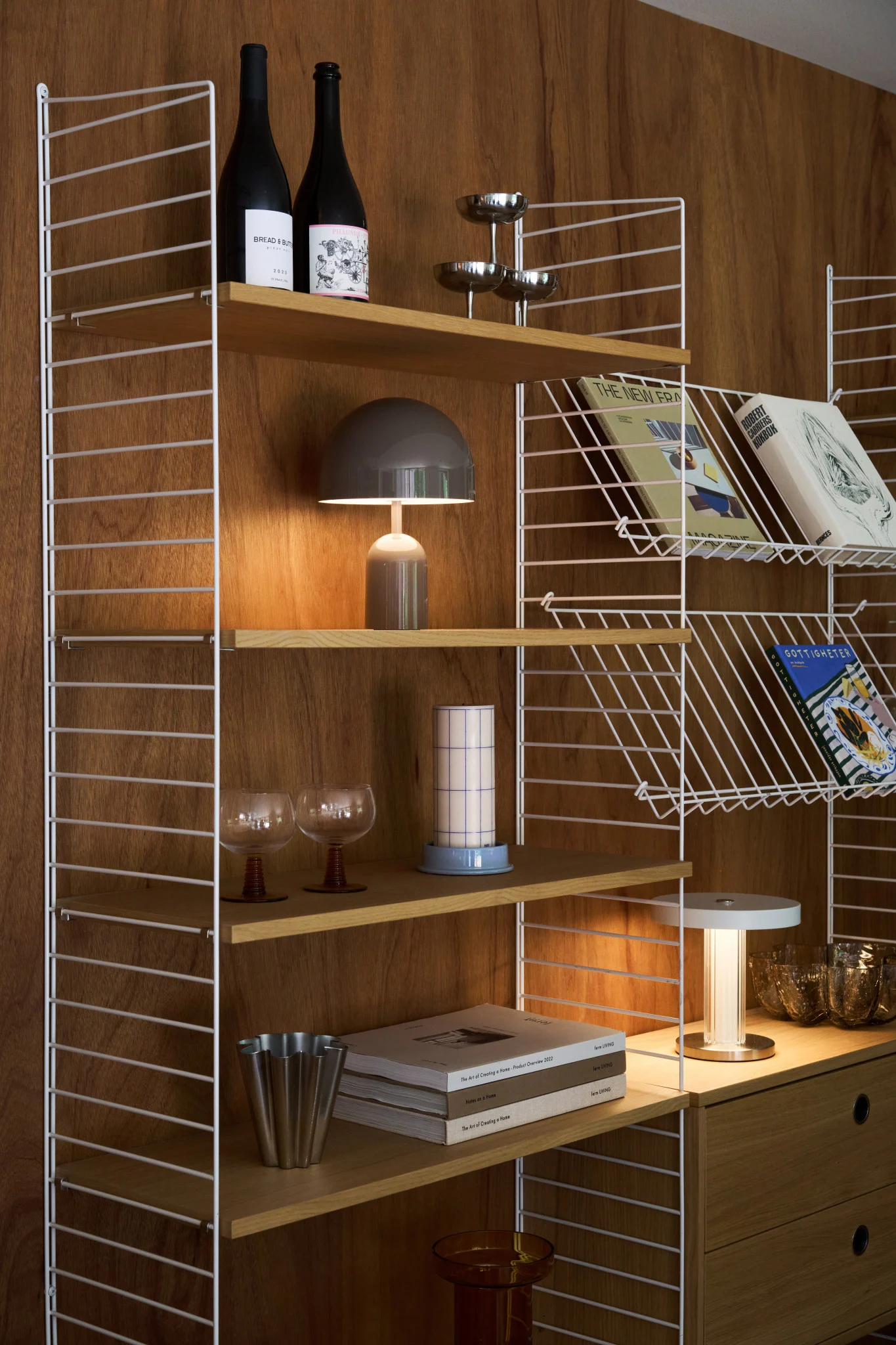 Ein stilvolles Holzregal mit weißen Drahtrahmen enthält Bücher, Flaschen, eine Pilzlampe und verschiedene Dekoartikel.