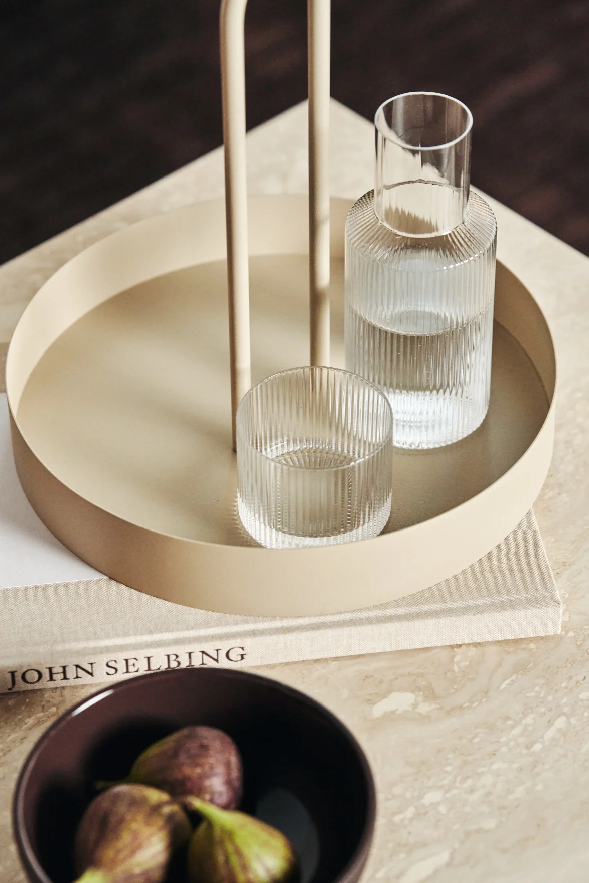 Auf einem beigefarbenen Tisch steht das beigefarbene Grib Tray Tablett, darauf ein Glas und die Karaffe aus der Ripple-Kollektion, alles von Ferm Living.
