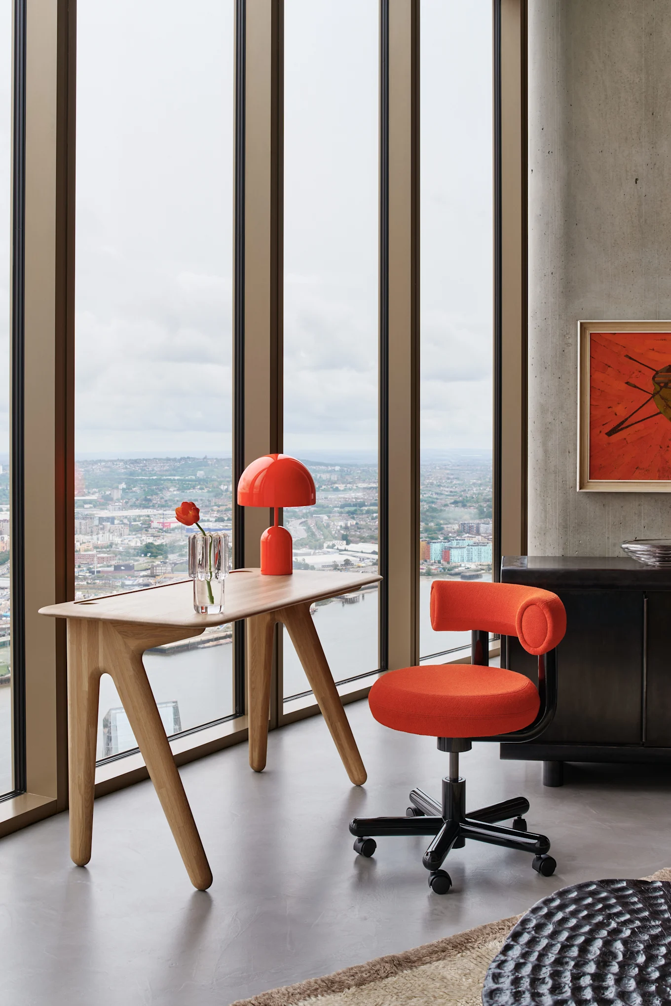 Ein modernes Büro mit einem orangefarbenen Drehstuhl und passender Lampe an einem hellen Holzschreibtisch mit Blick auf eine Stadt.