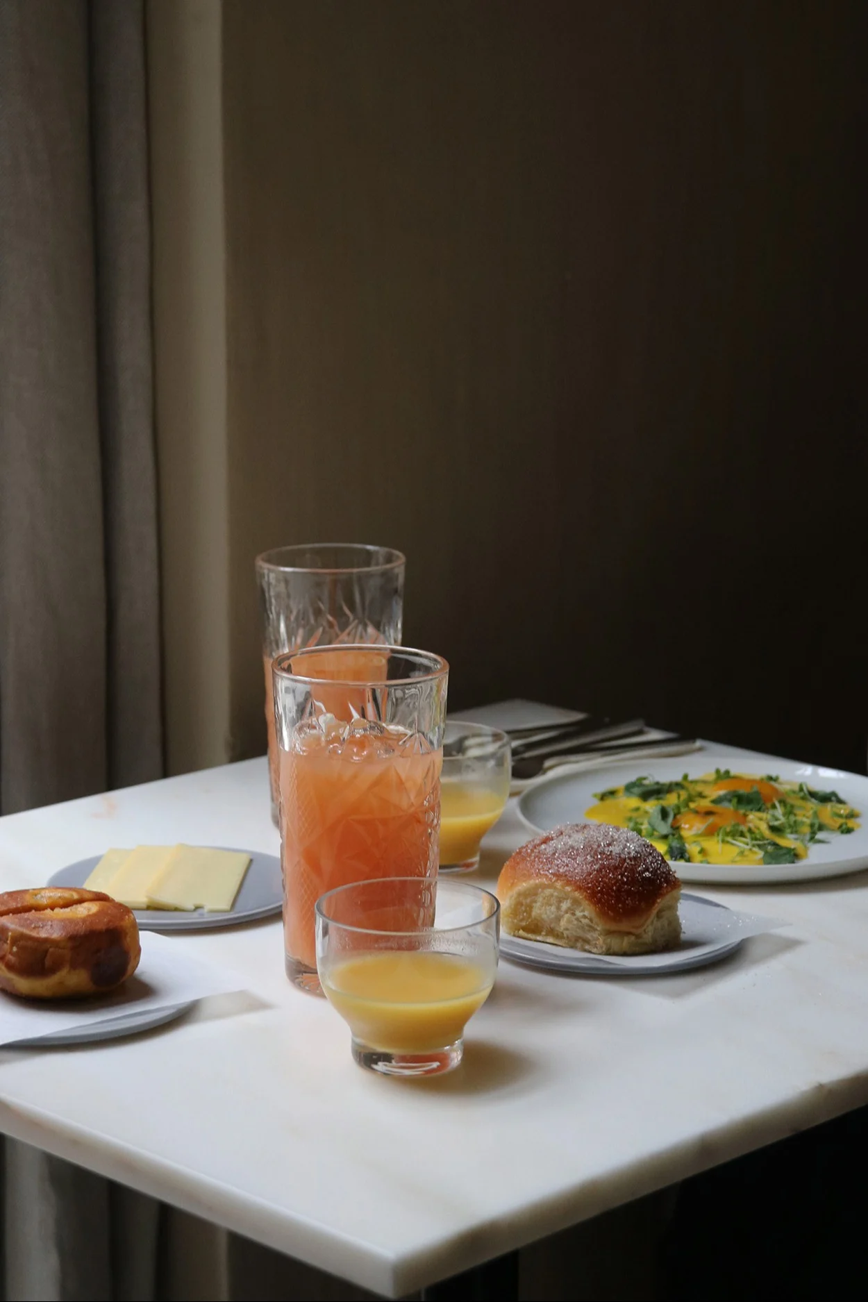 Ein Frühstück auf einem weißen Marmortisch mit Säften, einem herzhaften Brötchen, einem süßen Brötchen, Käsescheiben und Rührei mit Grünzeug.