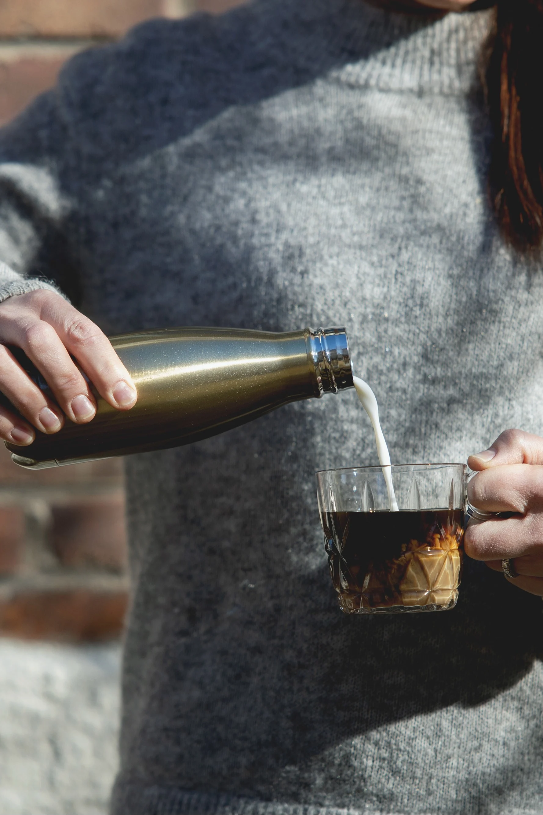 Person in grauem Pullover gießt Milch aus goldener Thermoskanne in ein Glas Eiskaffee.