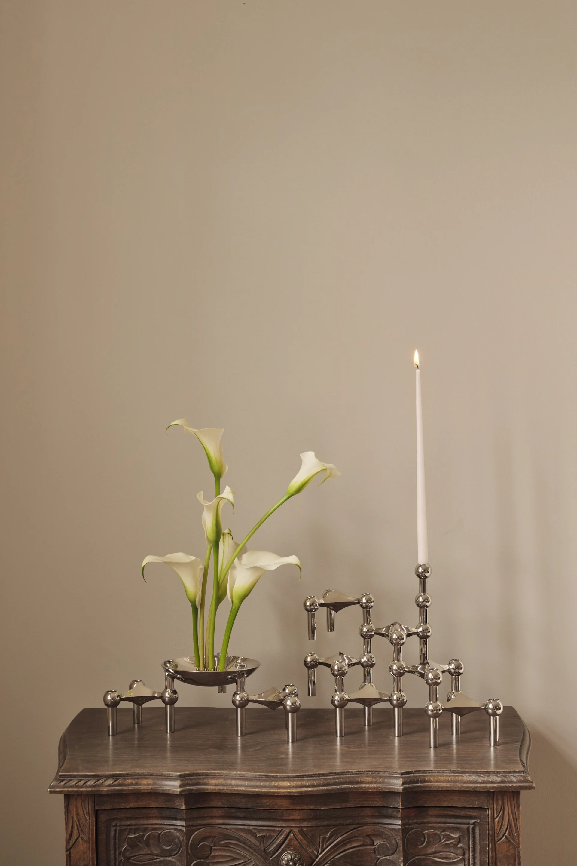 Weiße Calla-Lilien in einer Silberschale neben einem modularen silbernen Kerzenhalter mit einer brennenden weißen Kerze auf einem geschnitzten Holztisch.