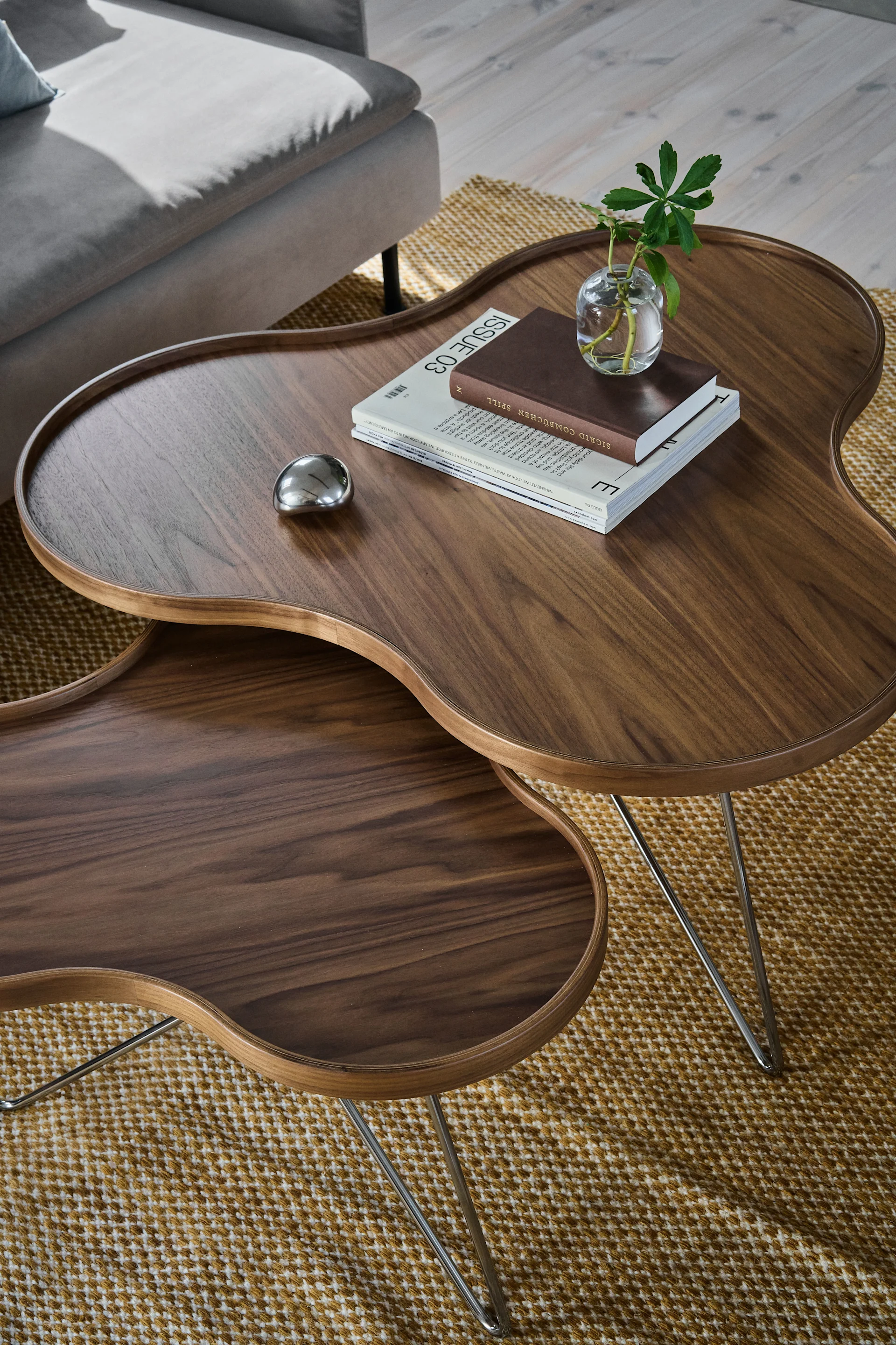 Zwei dunkle, nierenförmige Couchtische aus Holz auf einem gemusterten Teppich, mit einer Pflanze, Büchern und einem metallischen Objekt.