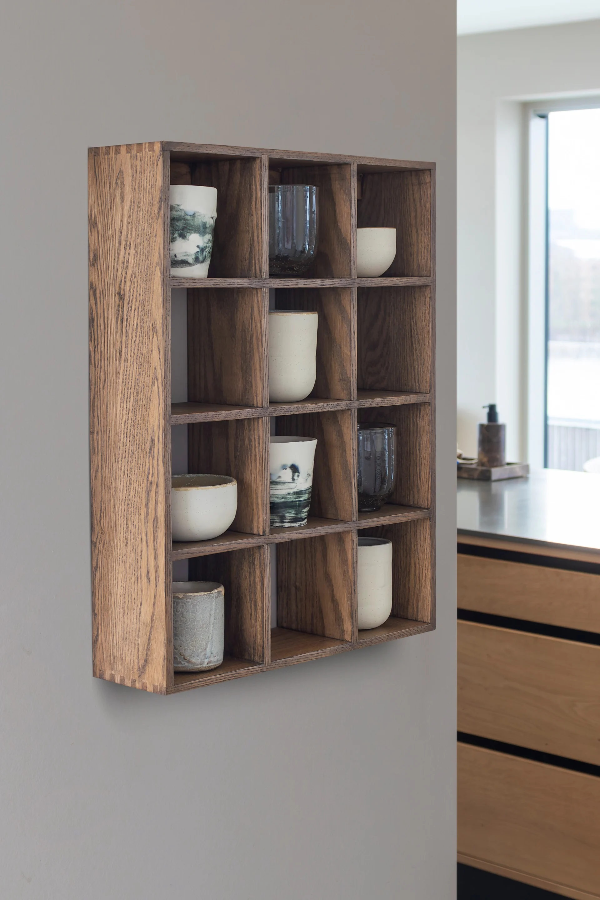 Ein wandmontiertes Holzregal mit zwölf quadratischen Fächern, das verschiedene Keramikbecher und -schalen präsentiert.