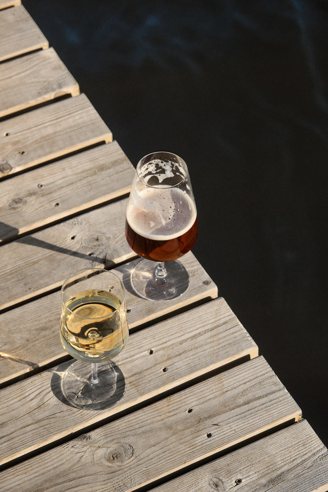 Zwei Gläser, Weißwein und Bier, auf einem Holzsteg neben dunklem Wasser im Sonnenlicht.
