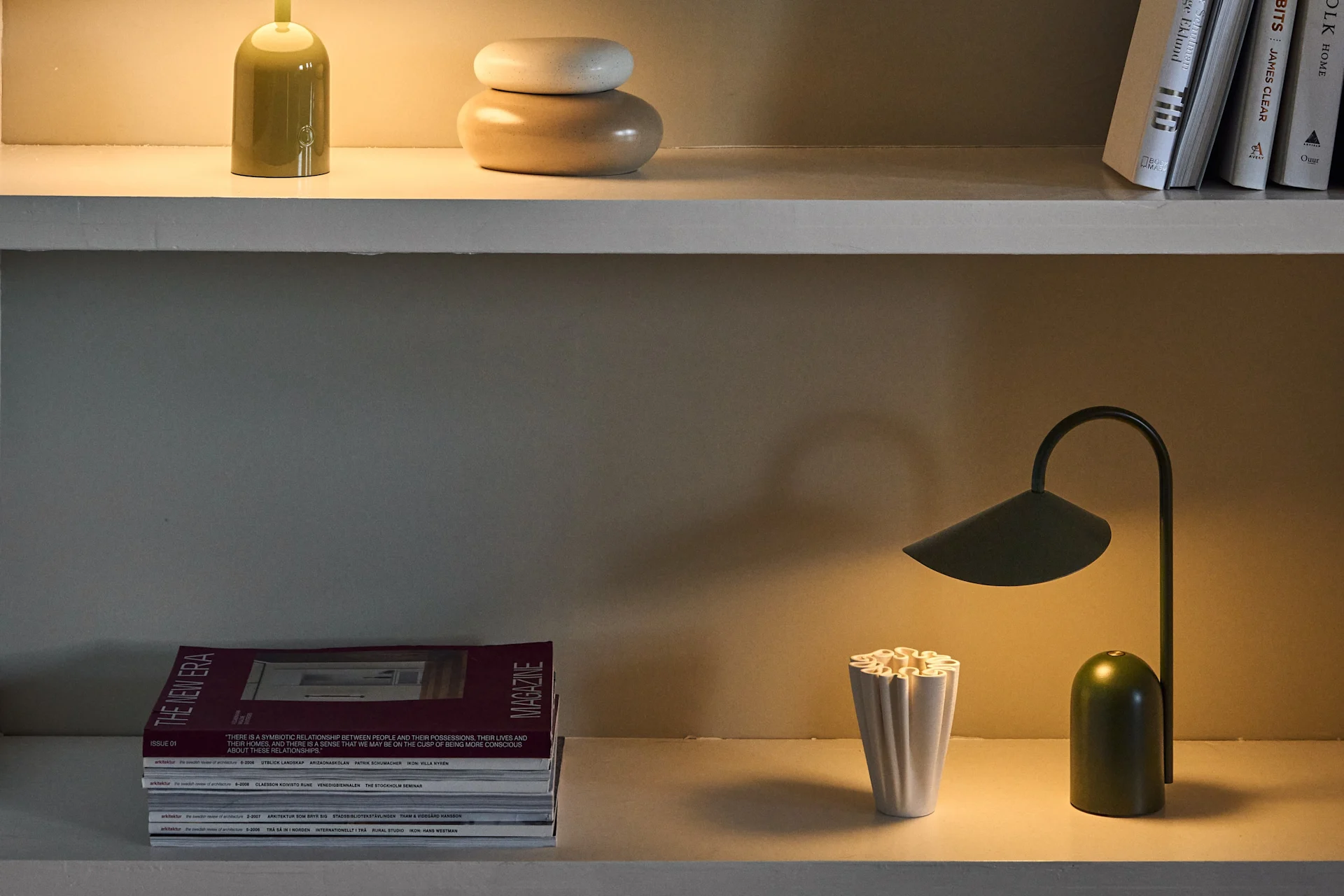 Drei Regale zeigen verschiedene Tischlampen in Grün und Lila, zusammen mit Büchern, Zeitschriften und Deko-Objekten.