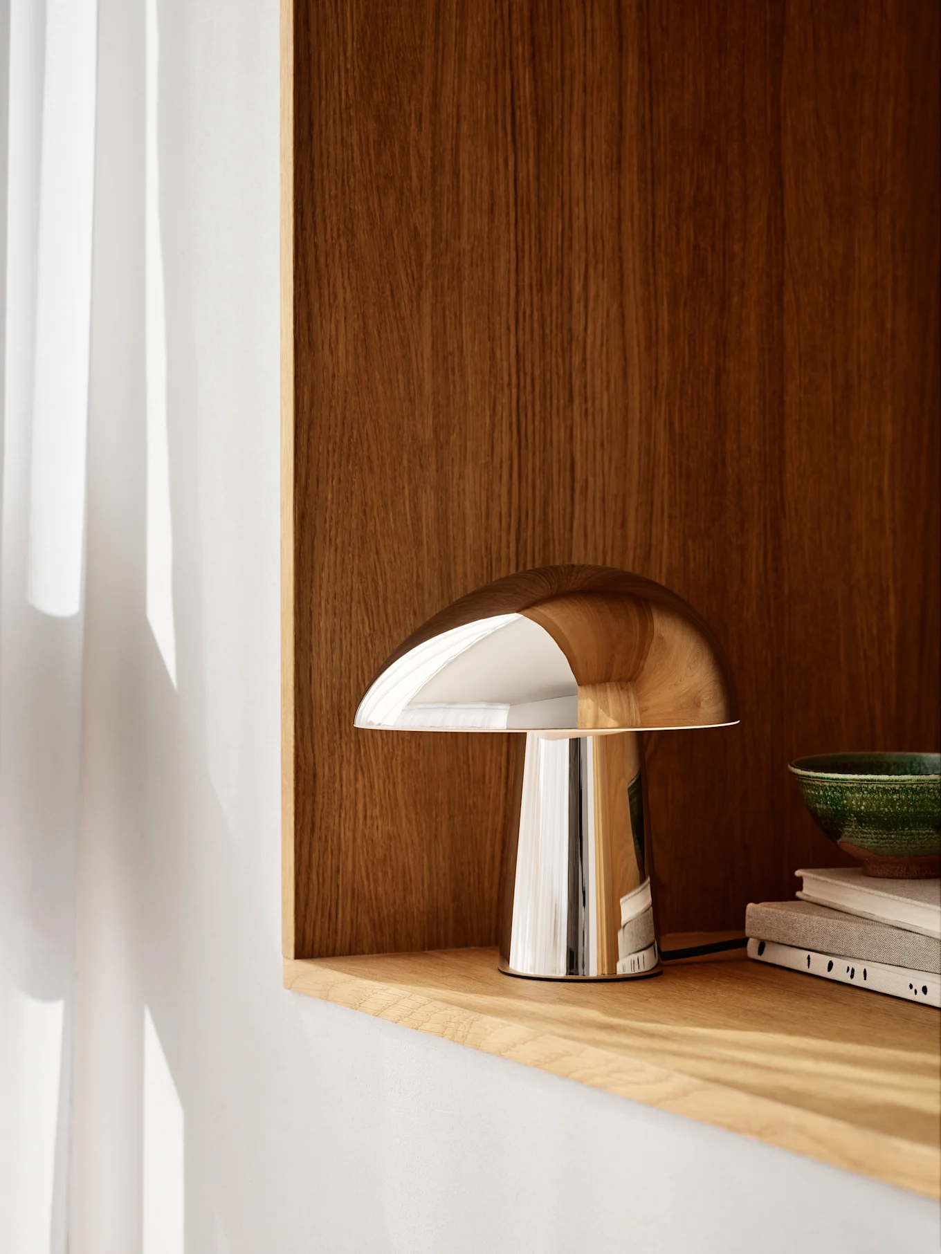 Eine polierte, verchromte, pilzförmige Lampe steht auf einem hellen Holzregal, dahinter eine dunkle Holzwand. Eine grüne Schale und Bücher sind rechts zu sehen.