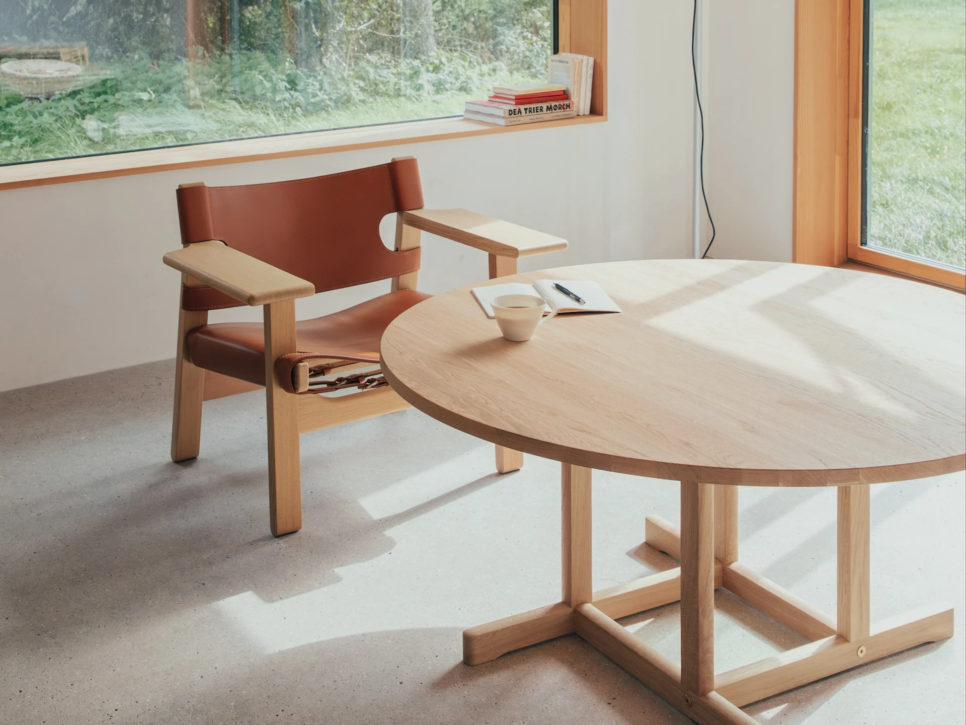 Helles Zimmer mit braunem Ledersessel, rundem Holztisch mit Tasse und Notizbuch, sonnenbeschienen von Fenster mit Blick ins Grüne.