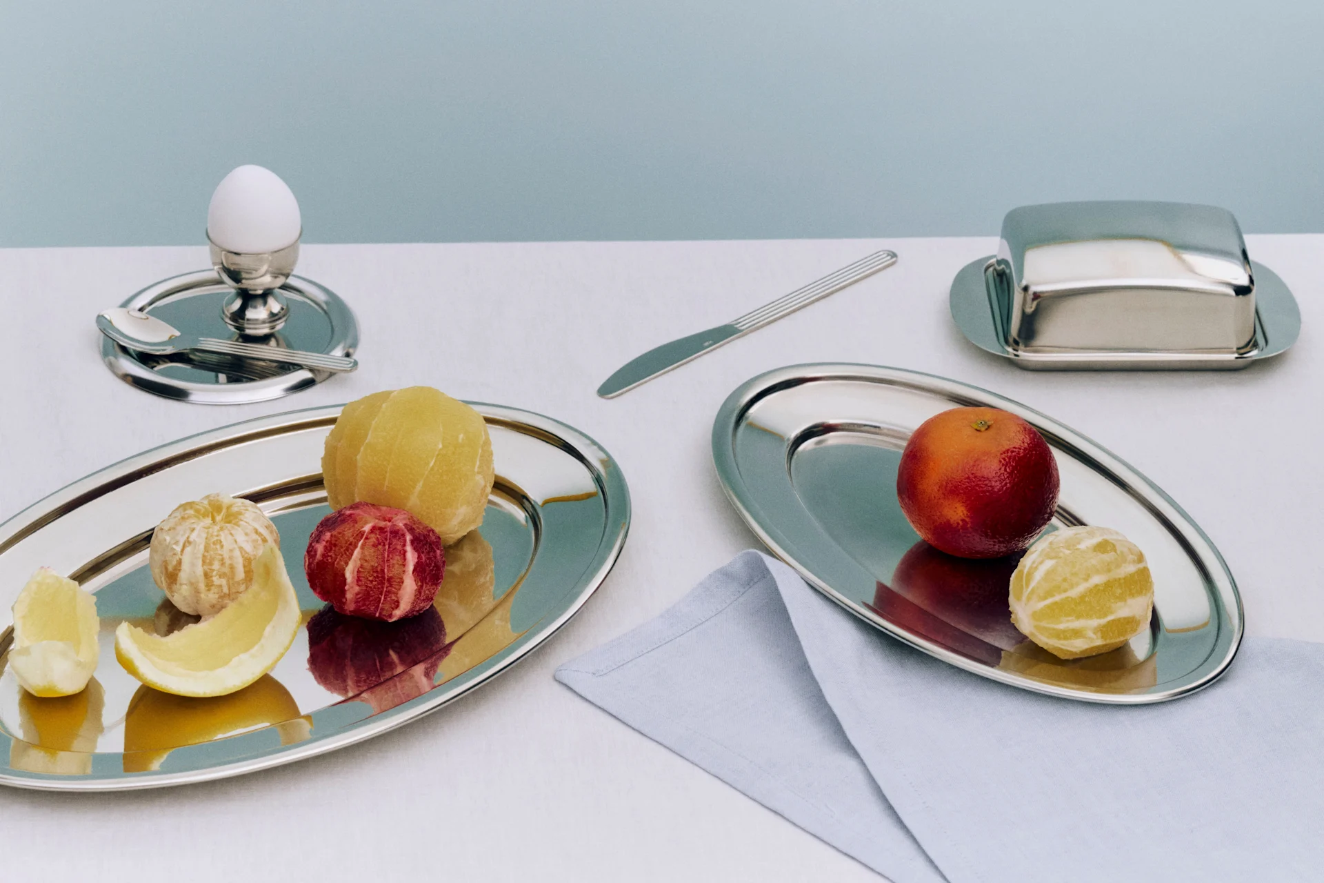 Frühstücksgedeck mit geschälten und ungeschälten Zitrusfrüchten auf Silbertabletts, Ei im Eierbecher und Butterdose.