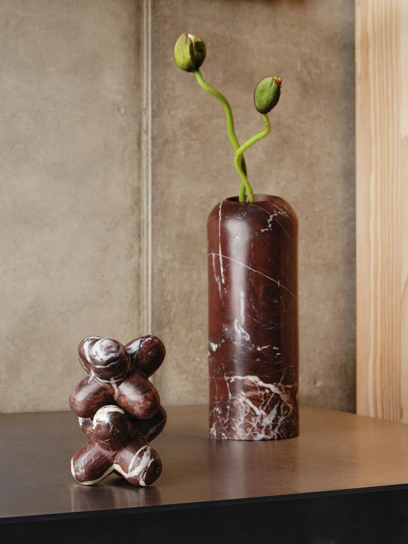 Zwei Marmorobjekte auf einem Regal: eine hohe dunkelrote Vase mit grünen Blumenknospen und eine abstrakte Skulptur.