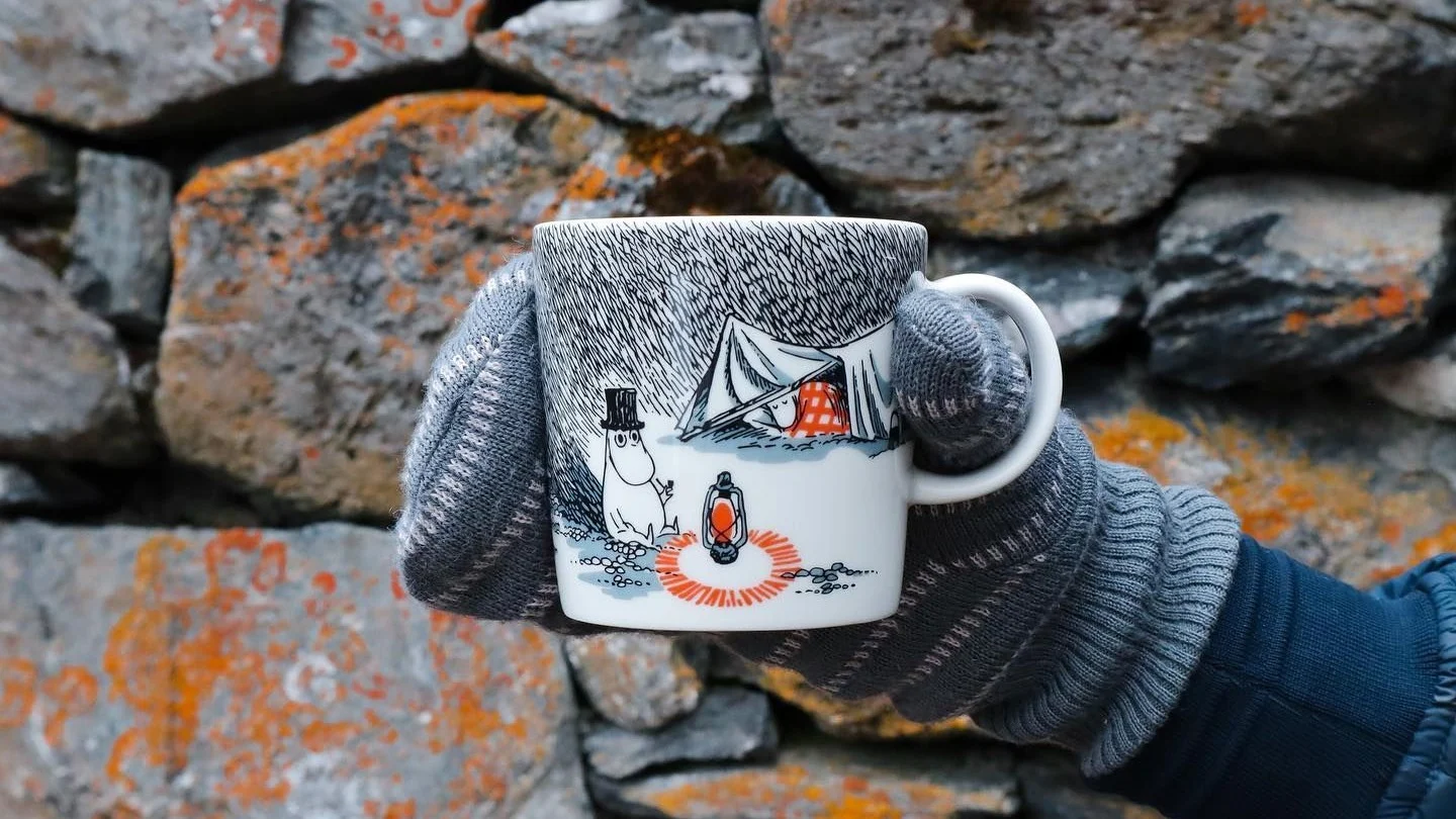 @moominbyingri hält die Schlaf gut-Mumin-Tasse vor einer Steinmauer in der Hand, die einen Handschuh trägt.