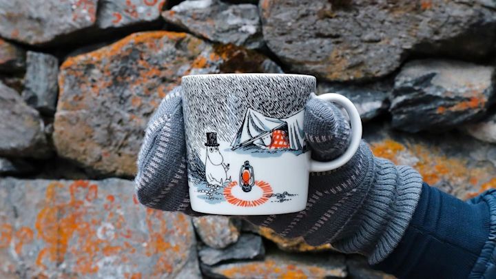 @moominbyingri hält die Schlaf gut-Mumin-Tasse vor einer Steinmauer in der Hand, die einen Handschuh trägt.