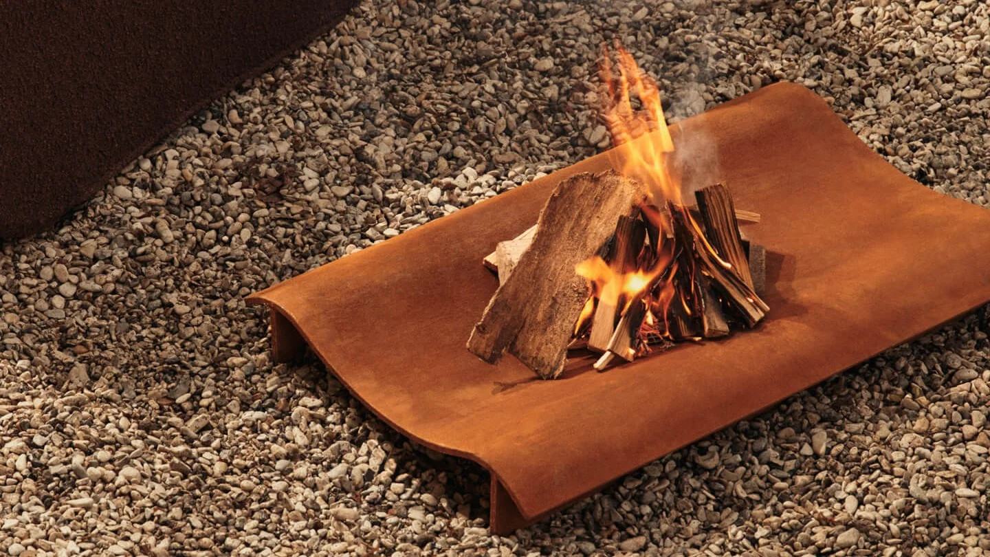 Kleines Lagerfeuer mit brennendem Holz auf einer rechteckigen, rostigen Metallschale über Kies.