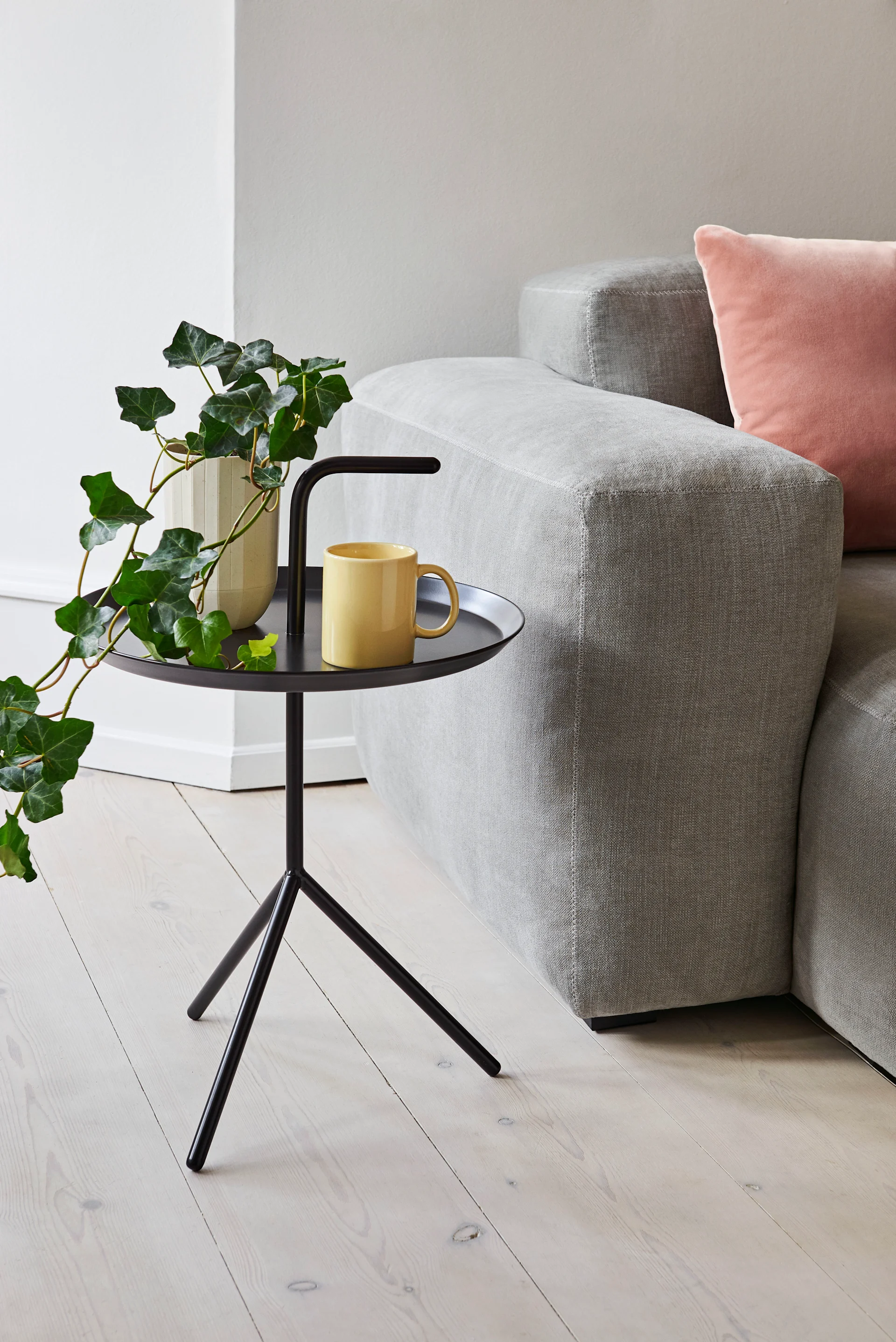 Ein schwarzer Stativ-Beistelltisch mit Efeupflanze und gelber Tasse, neben einem grauen Sofa.