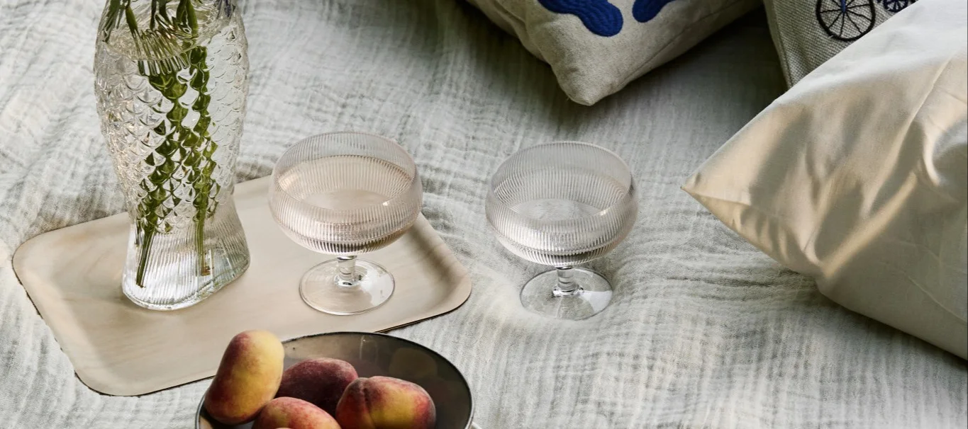 Picknick-Arrangement mit fischförmiger Vase, zwei geriffelten Gläsern und Pfirsichen auf einer hellen Decke.