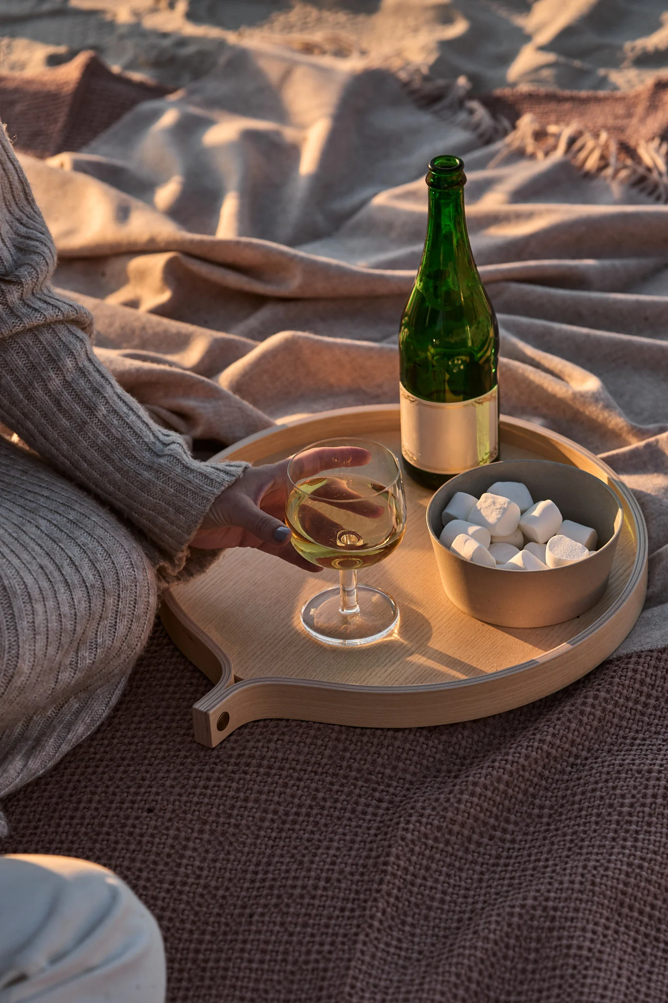 Person hält Weißwein auf einem Holztablett mit einer grünen Flasche und Marshmallows auf Decken während der goldenen Stunde.