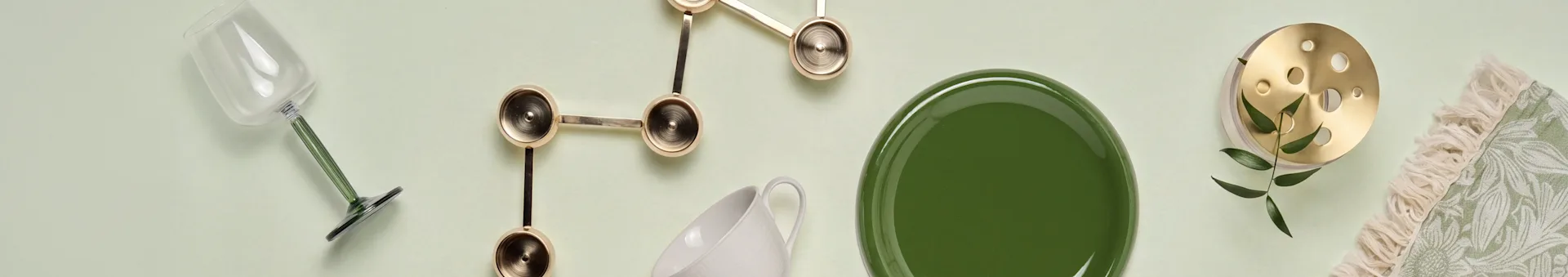 Flachansicht: Glas mit grünem Stiel, goldene Skulptur, weiße Tasse, grüner Teller, Messingobjekt mit Blättern und grünes gemustertes Textil mit Fransen.