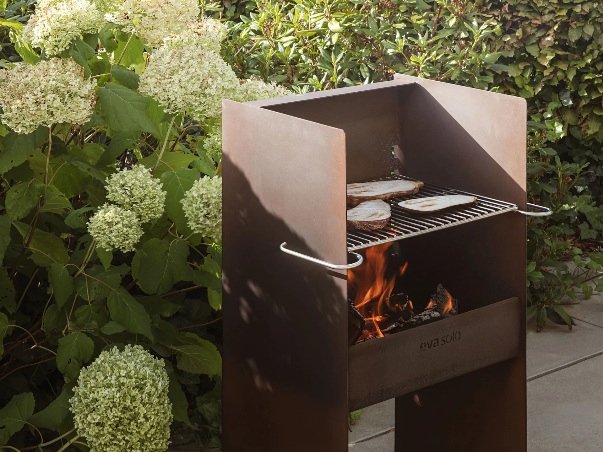 Ein rustikaler Metall-Außengrill mit Flammen und Auberginenscheiben, die auf dem Rost garen.