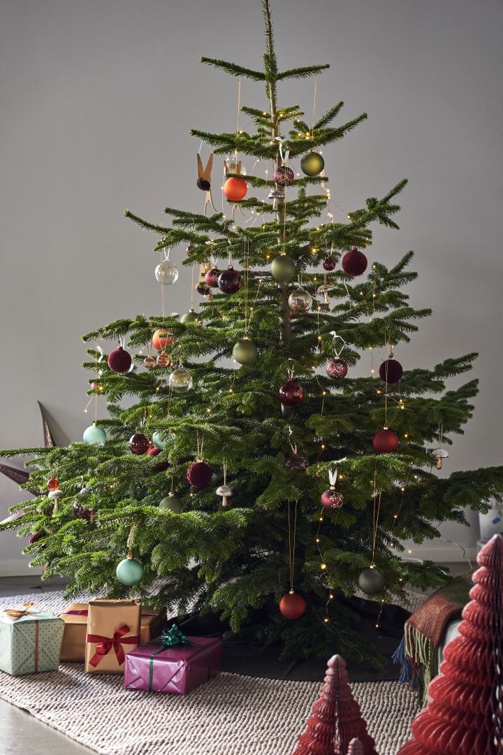 Dekorieren Sie Ihren Baum dieses Jahr mit dem Weihnachtsbaumschmuck 2021 in den 4 verschiedenen Stilen der Nest Trends – Nurture, Share, Boost und Cultivate. Hier sehen Sie einen prachtvollen Weihnachtsbaum auf dem Flock Wollteppich von Scandi Living in Beige, dekoriert mit Schmuck in satten Rot-, Orange- und Grüntönen. 