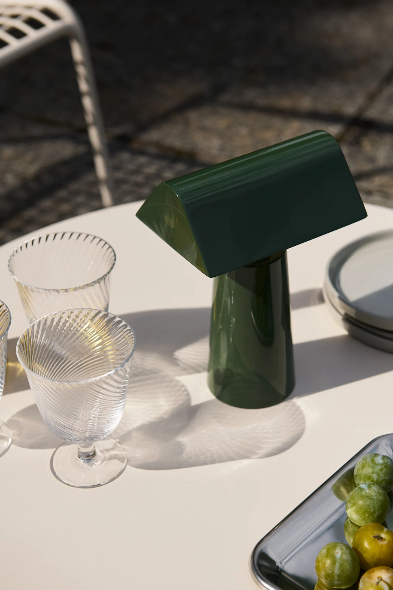 Eine dunkelgrüne Tischlampe und zwei gerippte Gläser auf einem Gartentisch mit Pflaumen auf einem Tablett, im Sonnenlicht.