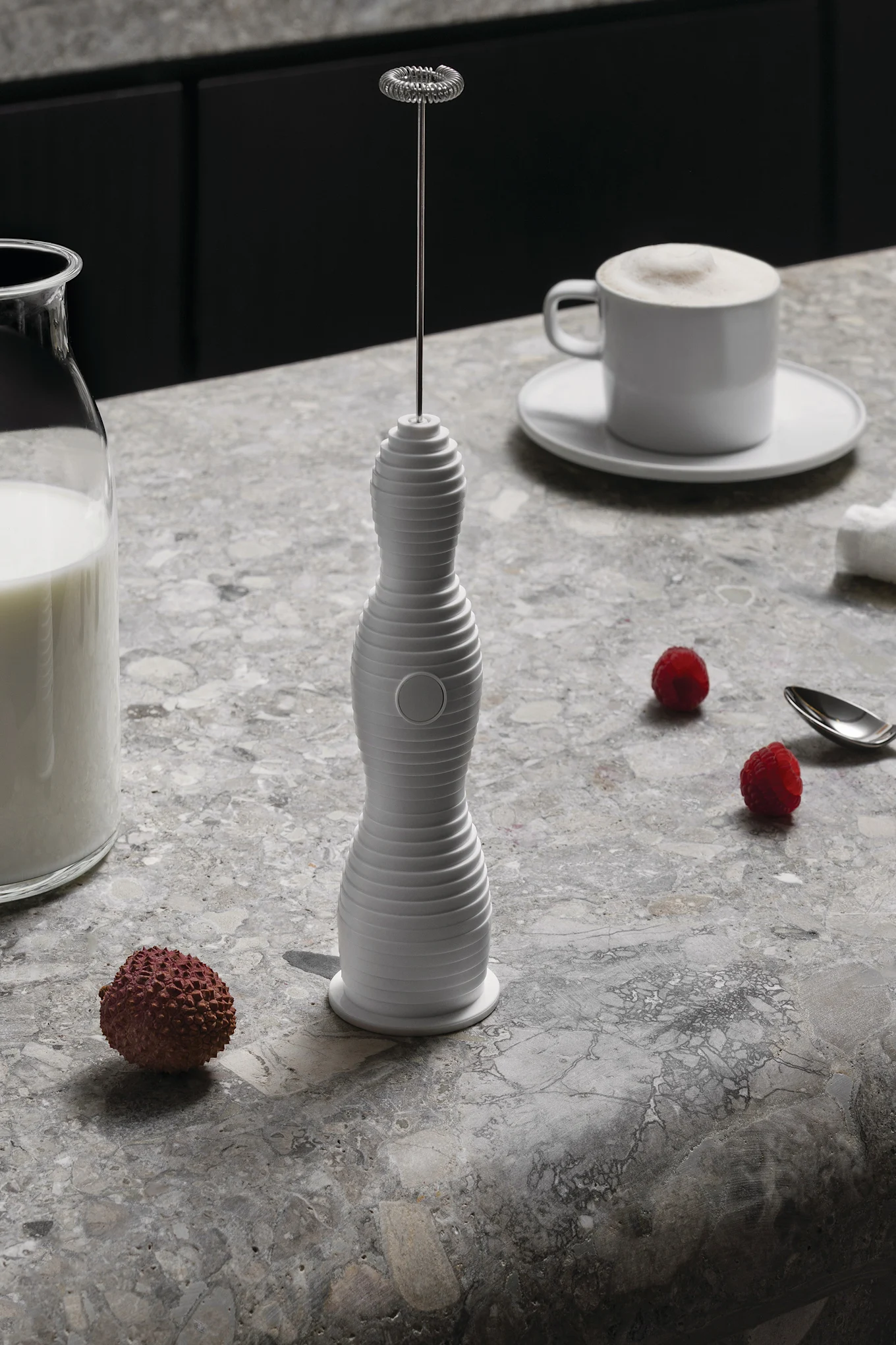 Ein weißer Milchaufschäumer steht auf einer marmorierten Theke neben einer Milchflasche, einer Tasse geschäumten Kaffees, Himbeeren und einer Litschi.
