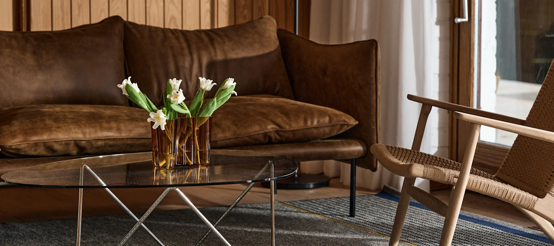 Modernes Wohnzimmer mit braunem Ledersofa, Glas-Couchtisch mit weißen Tulpen und Holzstuhl mit geflochtener Sitzfläche.