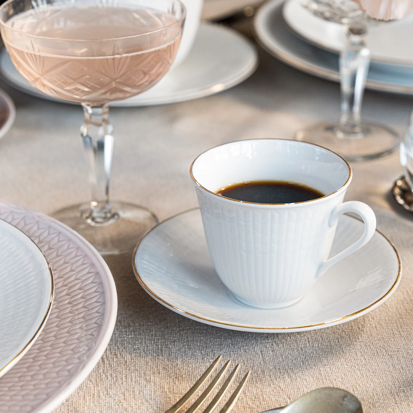Eine weiße Kaffeetasse mit Goldrand auf einer Untertasse, neben einem rosa Teller und einem Roséglas auf einem Leinentisch.