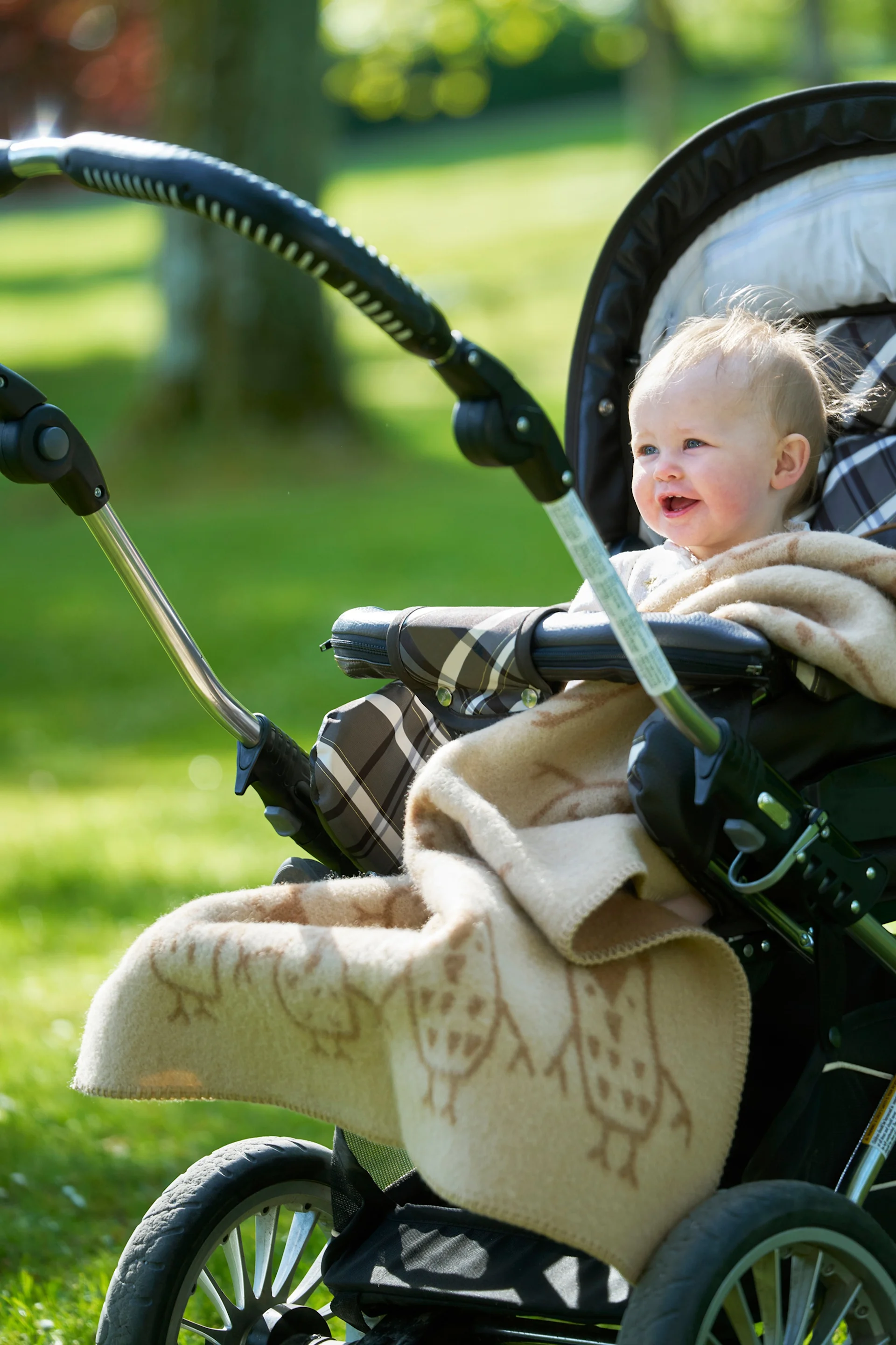 Ein lächelndes Baby mit hellem Haar, eingewickelt in eine Decke mit Eulenmuster, sitzt draußen in einem Kinderwagen in einem sonnigen Park.