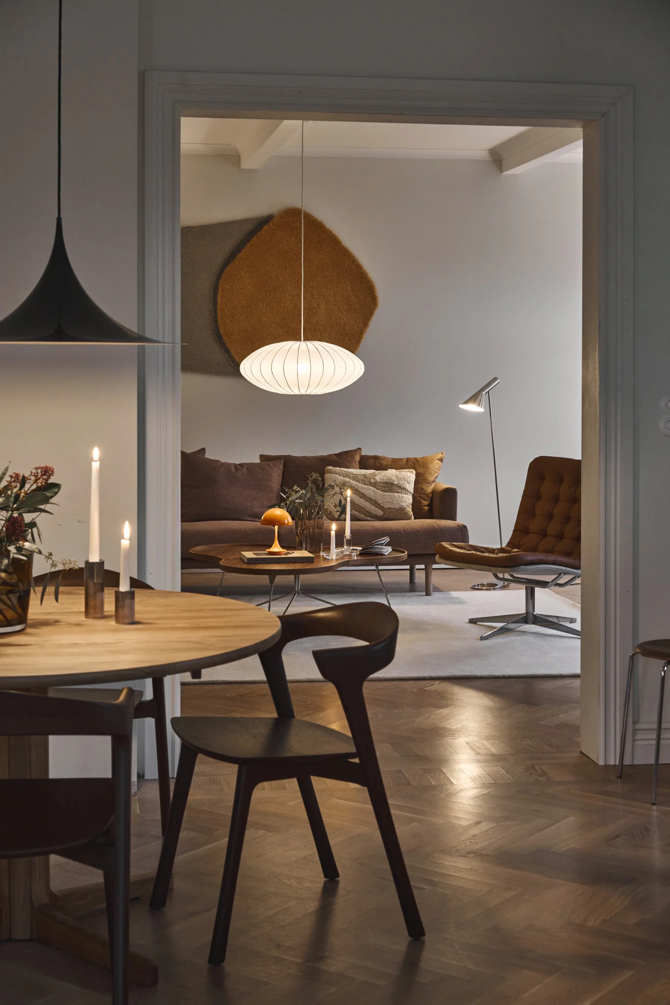 Blick von einem Esszimmer in ein Wohnzimmer mit braunem Sofa, Ledersessel und verschiedenen Lampen, darunter eine weiße ovale Pendelleuchte.