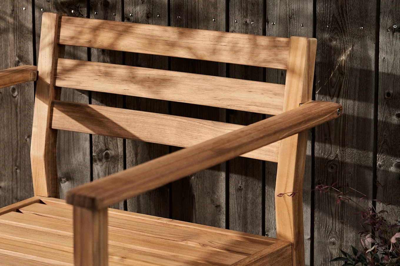 Gestreifter Holzstuhl mit Armlehnen vor einem dunklen Holzzaun, bedeckt mit Blattschatten.