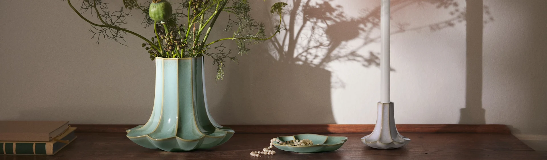 Eine geriffelte türkisfarbene Vase mit Blattwerk, eine Perlenkette in einer passenden Schale, eine weiße Kerze in einem geriffelten Halter und Bücher auf einer Holzoberfläche.