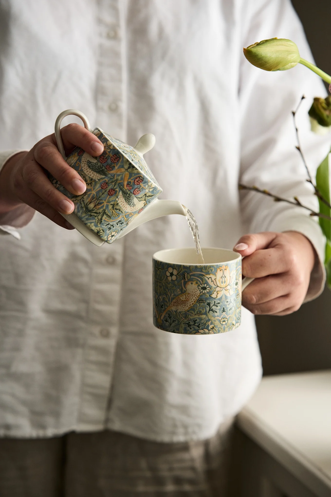 Hände gießen aus einer blauen Teekanne mit Vogel- und Beerenmuster in eine passende Tasse.